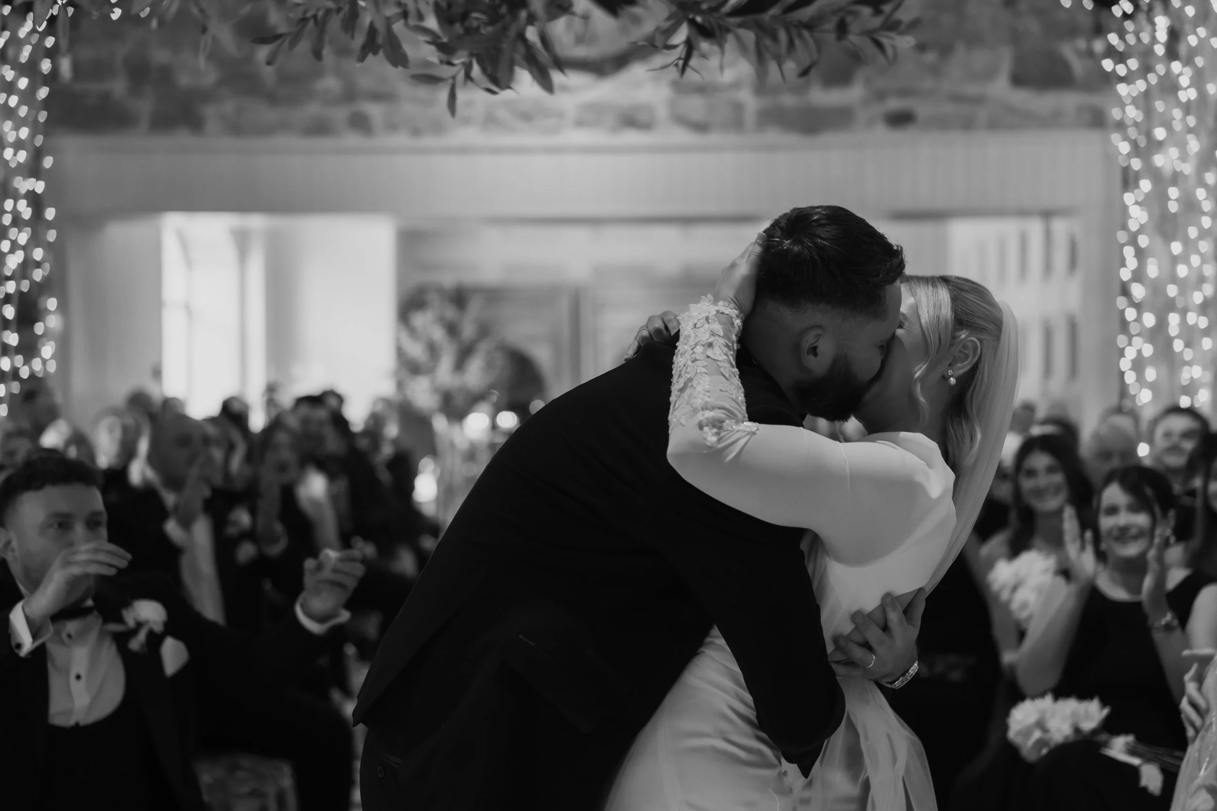 A couple sharing a kiss at a wedding reception, with guests watching and smiling in the background, decorated with string lights and greenery.