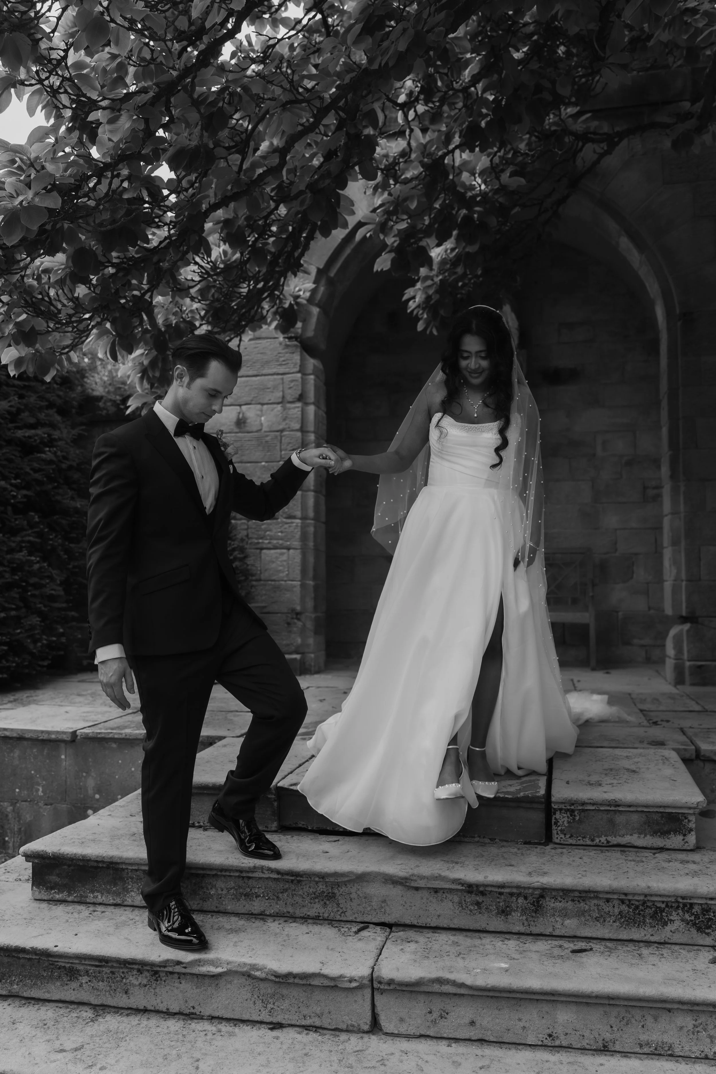 A black and white photo of a bride and groom holding hands and descending stone steps outside, with a brick archway and overhanging tree branches in the background. The groom is in a tuxedo and the bride in a wedding dress with a veil.
