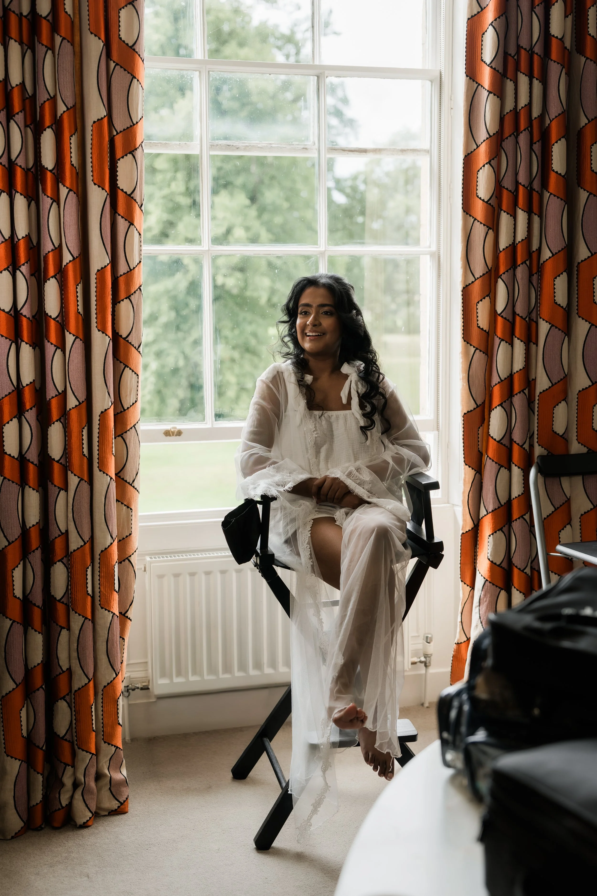 A woman sitting on a director's chair in front of a window with orange patterned curtains, smiling and wearing a white sheer dress, with lush greenery visible outside.