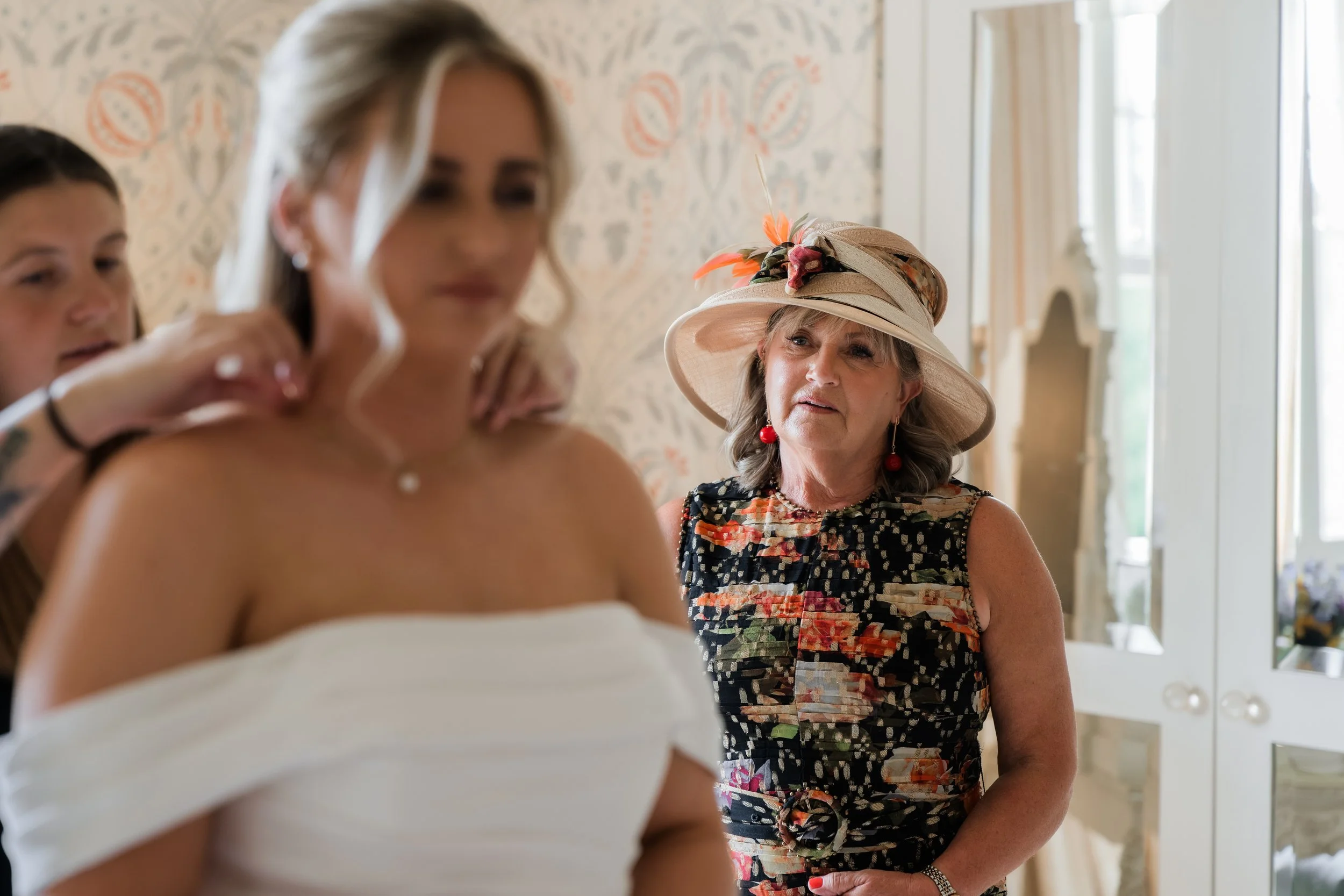 An elderly woman in a floral dress and large hat with feathers, earrings, and jewelry observing a woman getting ready, possibly at a wedding or special event, in a bright, decorated room.