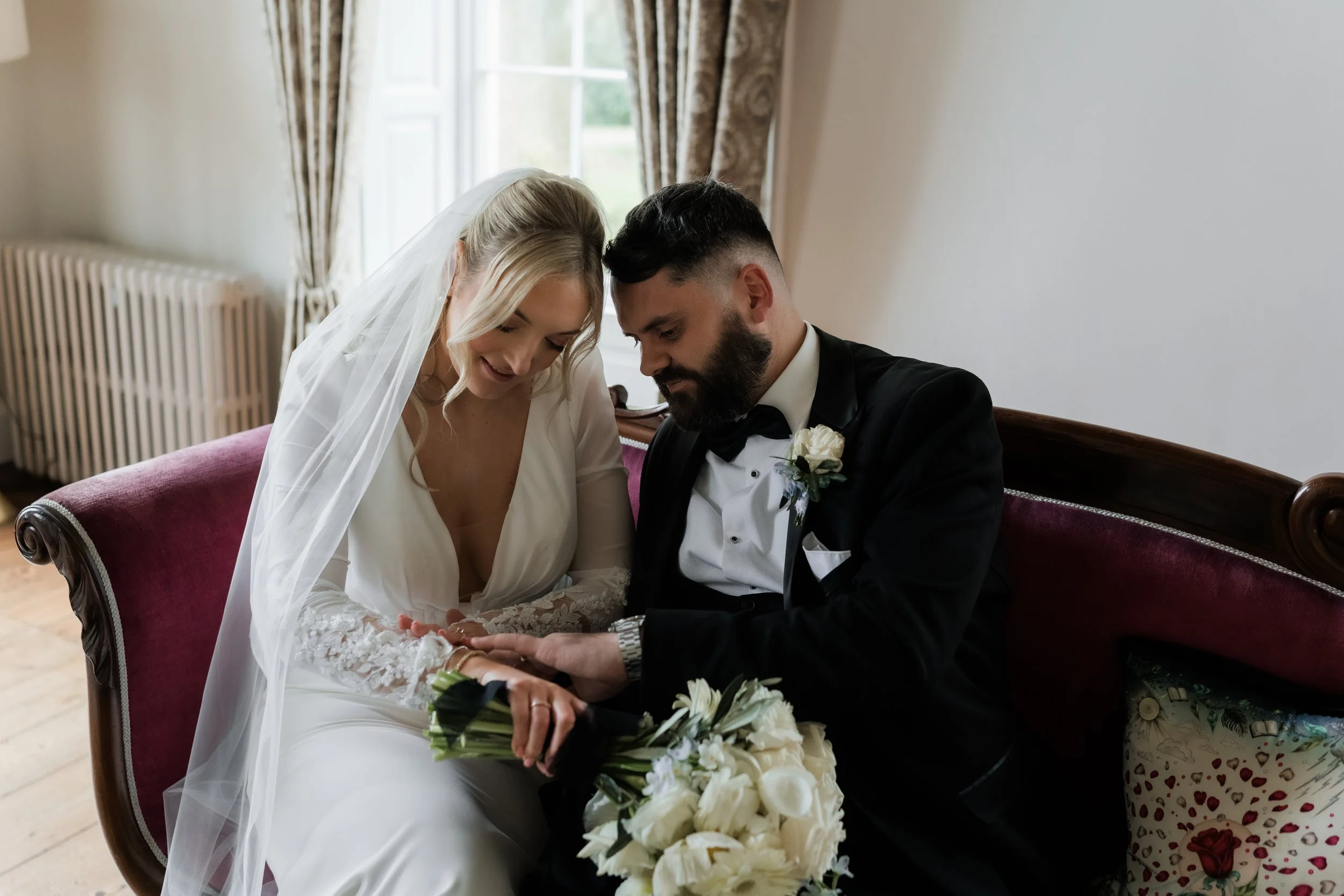 A bride and groom sitting on a vintage red velvet sofa, looking at each other's hands during their wedding ceremony.