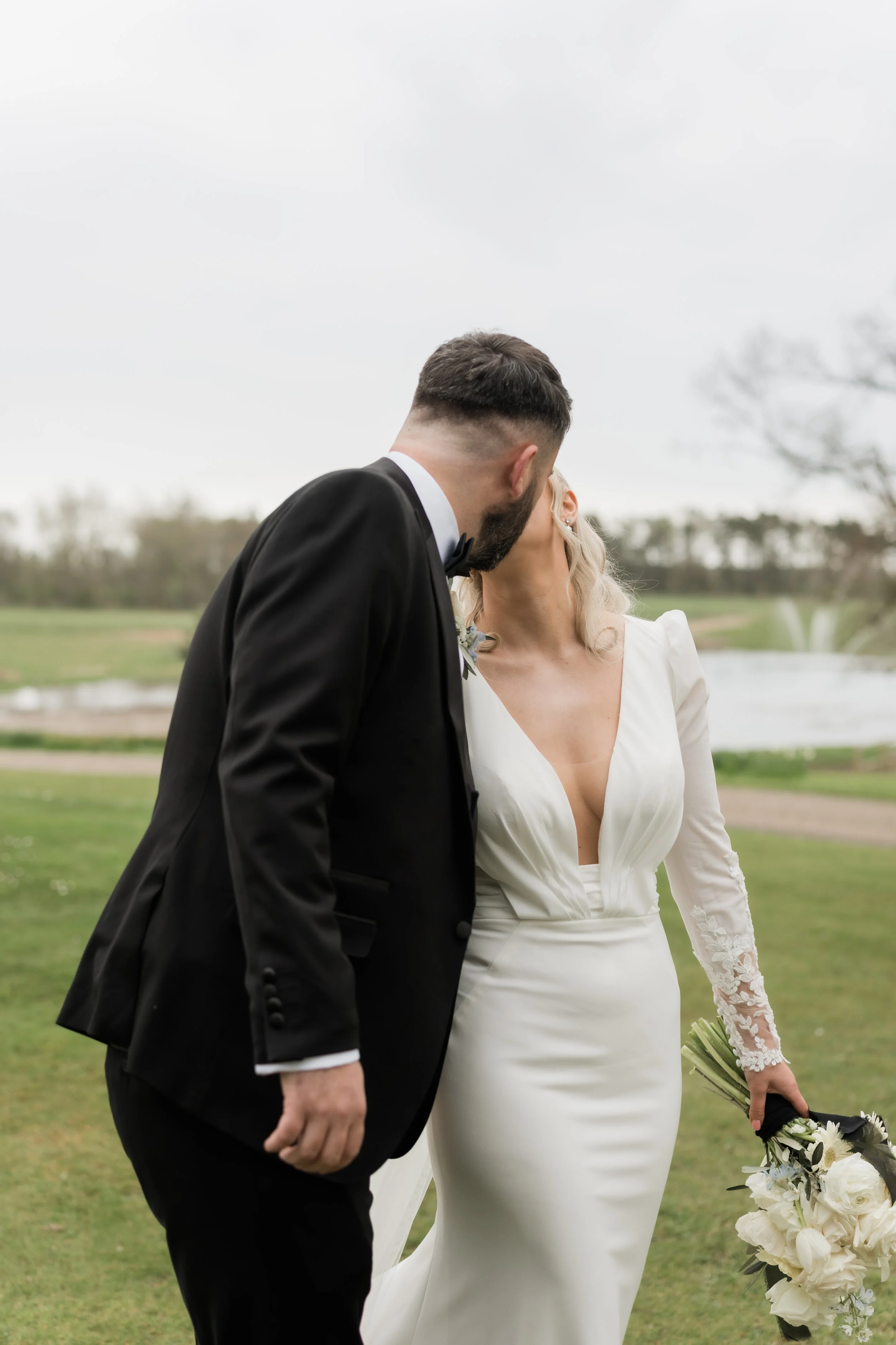 A bride and groom sharing a kiss outdoors on a cloudy day, with the bride holding a bouquet of white flowers.
