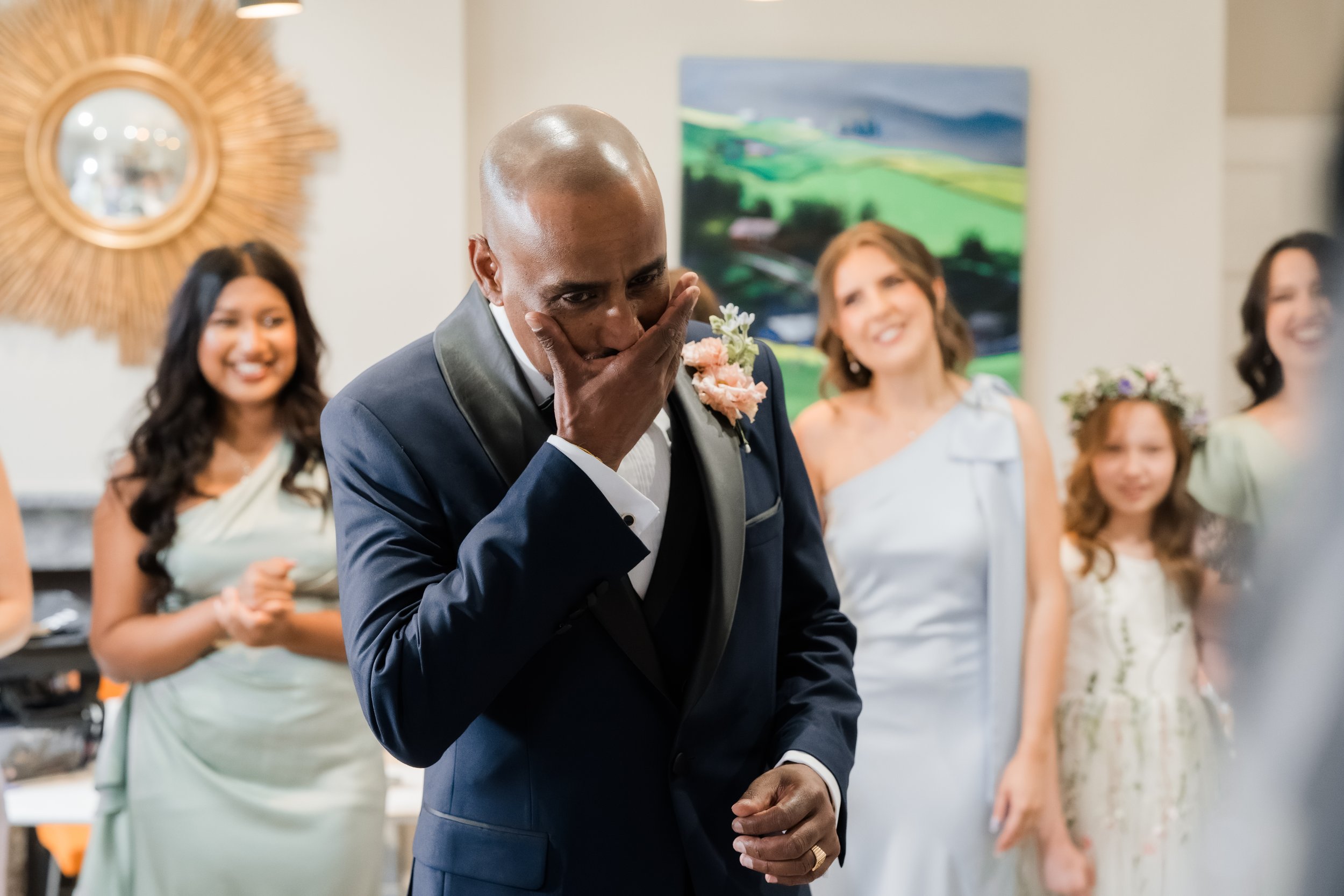 A man in a dark suit with a boutonniere covers his mouth with his hand while surrounded by women at a celebration or wedding.
