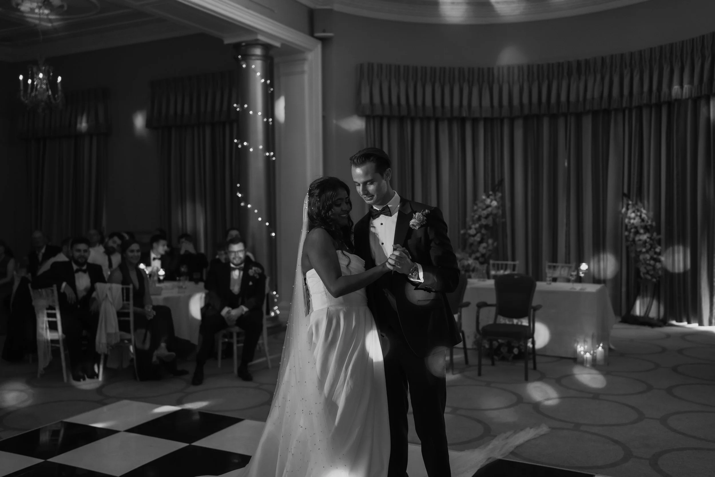 Black and white photograph of a bride and groom dancing at their wedding reception, surrounded by seated guests and elegant decor.