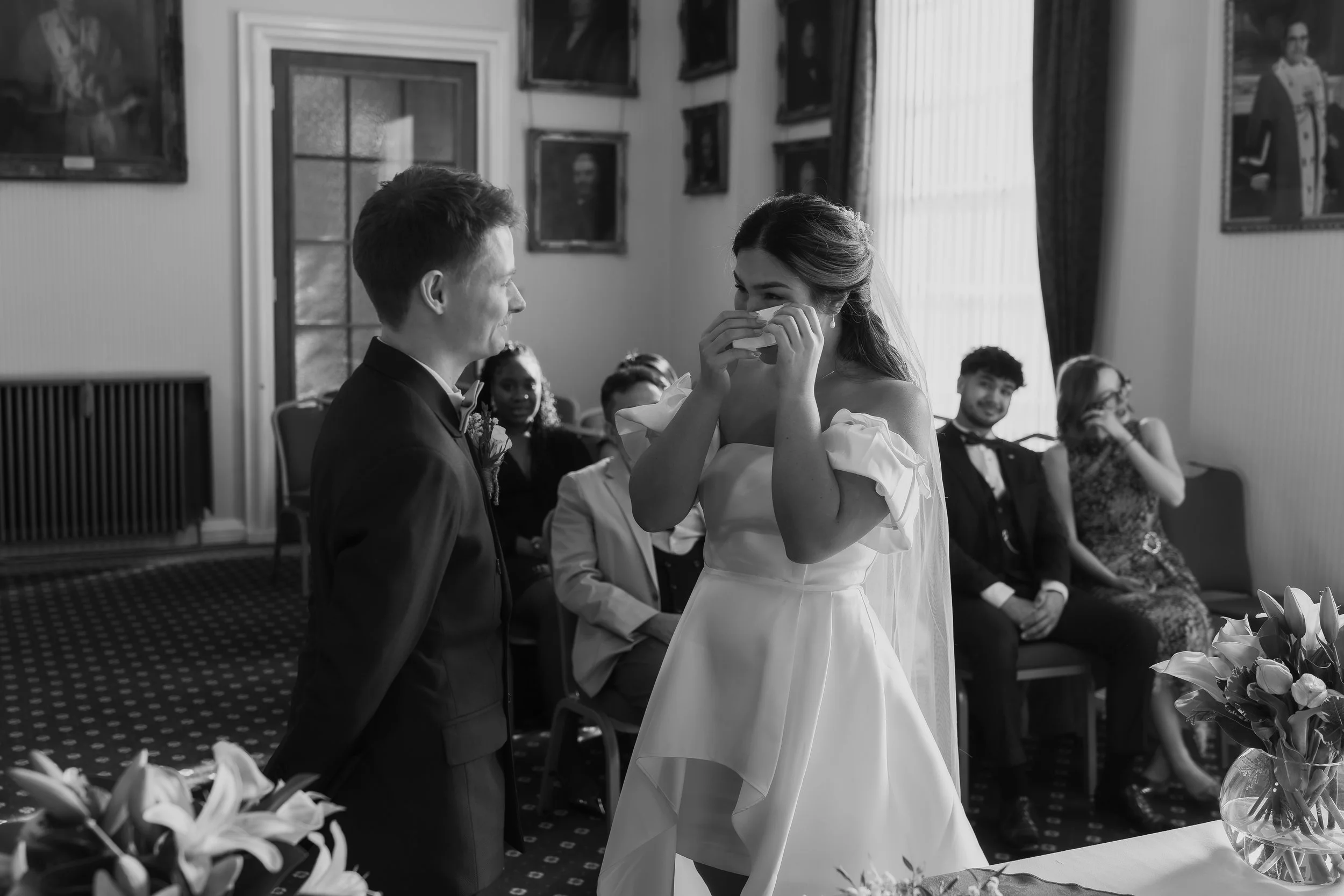 A black and white photo of a wedding scene where a woman in a white dress is emotional, holding a tissue near her face, standing opposite a man in a suit. There are guests seated in the background, watching the moment, with some smiling and others covering their faces, in a decorated room with framed pictures and floral arrangements.