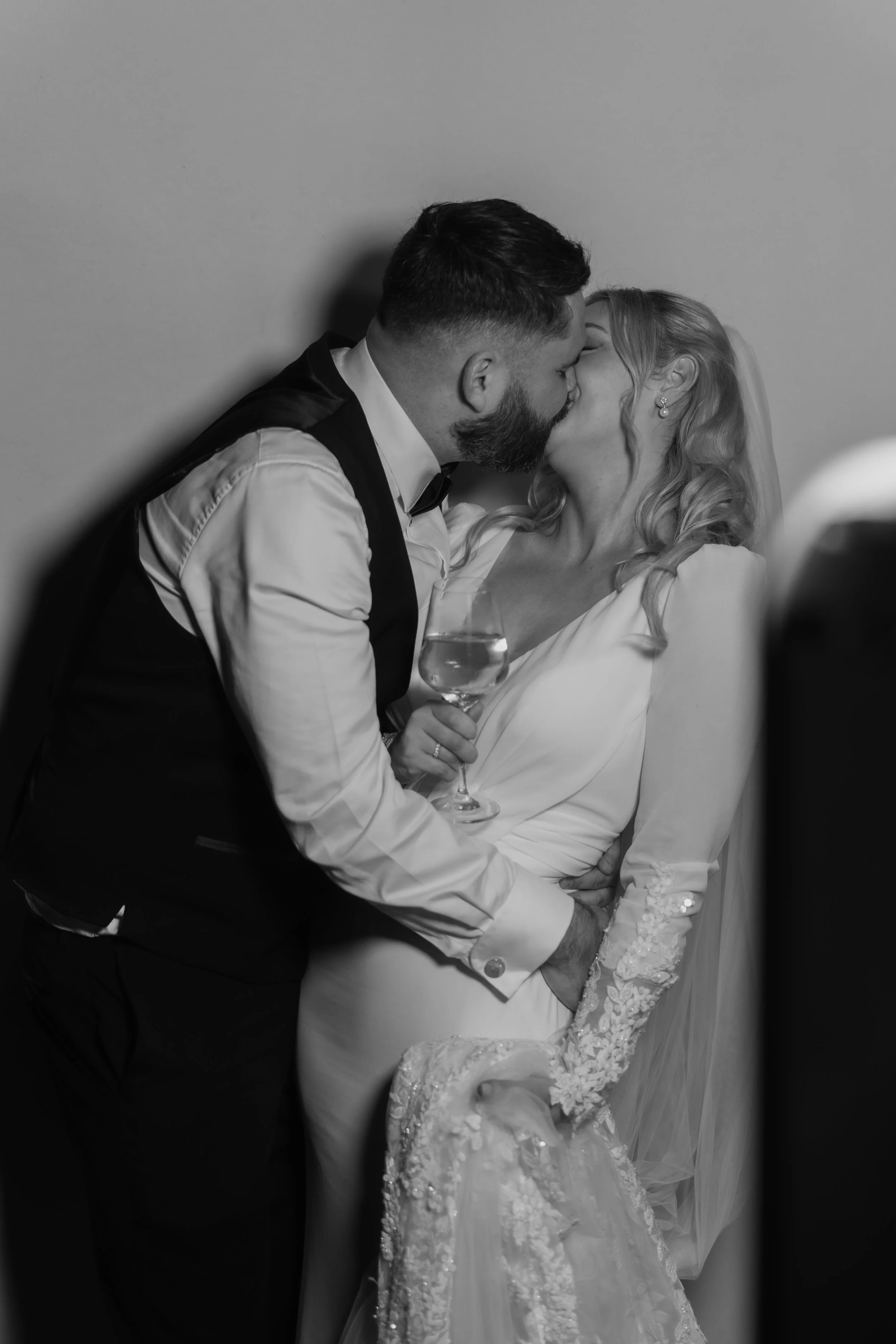 A black and white photo of a couple sharing a passionate kiss, with the man holding the woman who is holding a glass of wine, dressed in wedding attire.