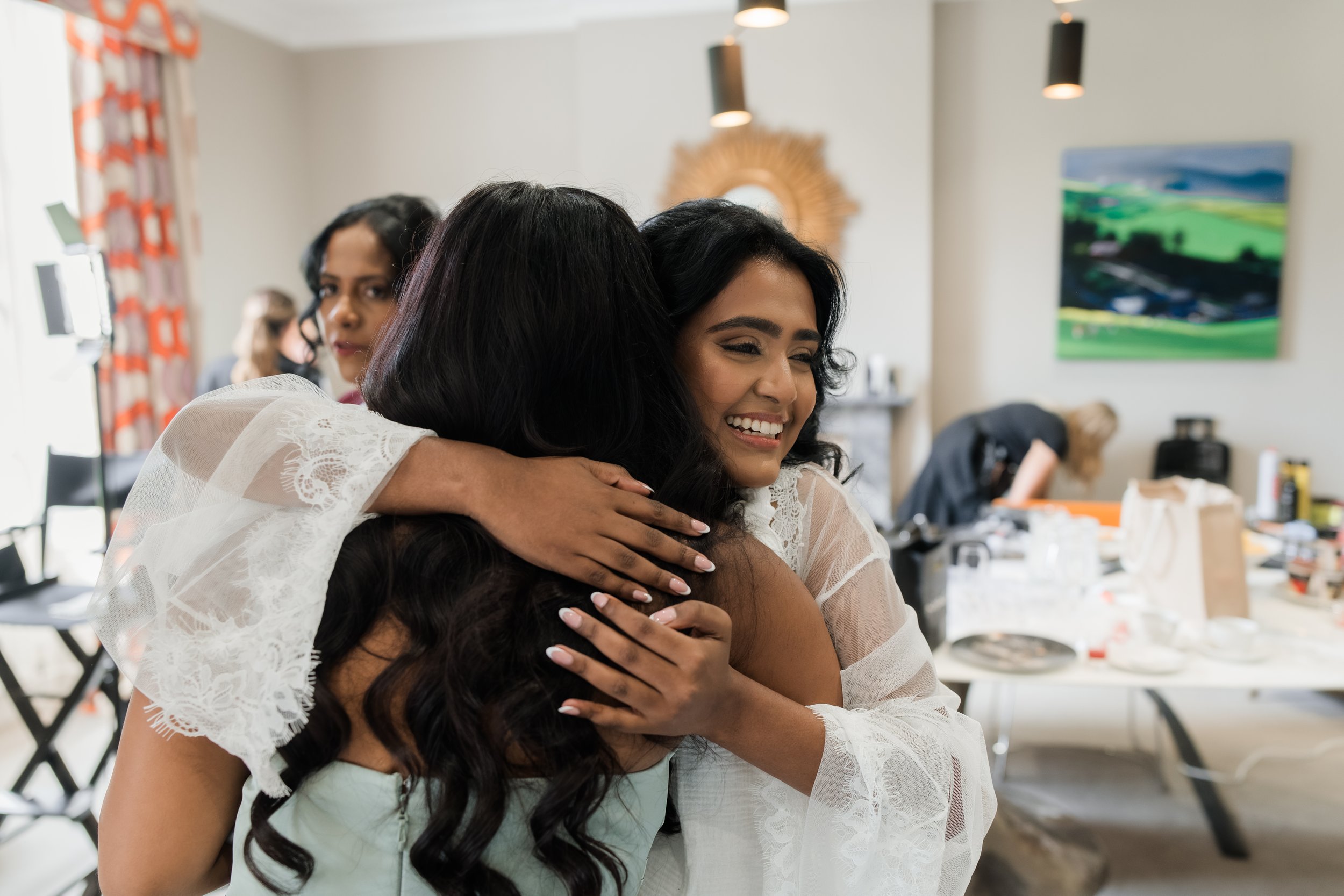 Two women hugging, one smiling, in a bright room with people and makeup in the background.