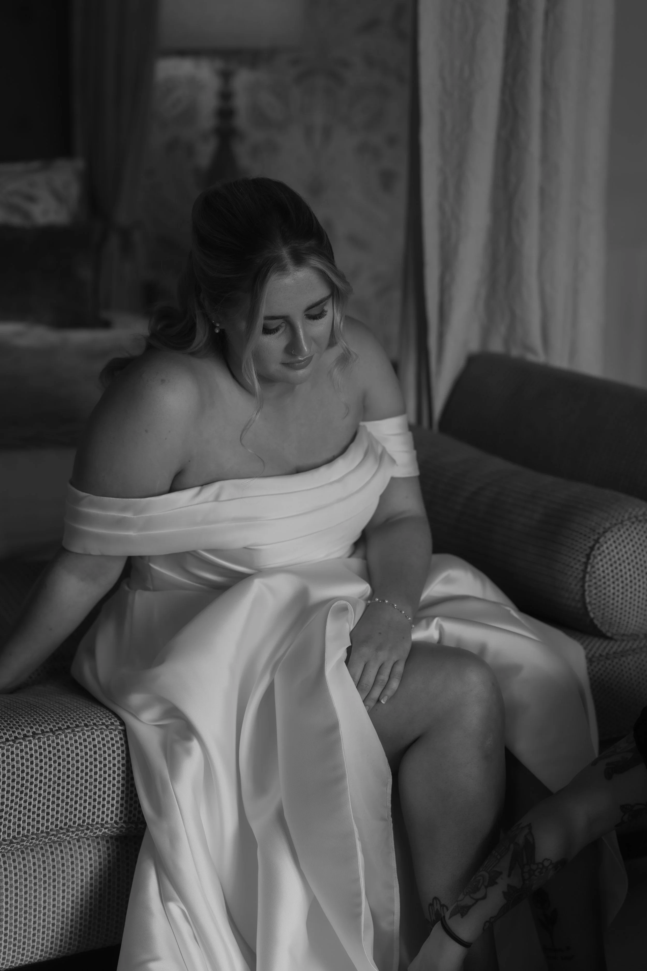 A black and white photo of a woman in a wedding dress sitting on a sofa, looking down. She has styled hair and earrings, and her dress is off the shoulder with pleated details.