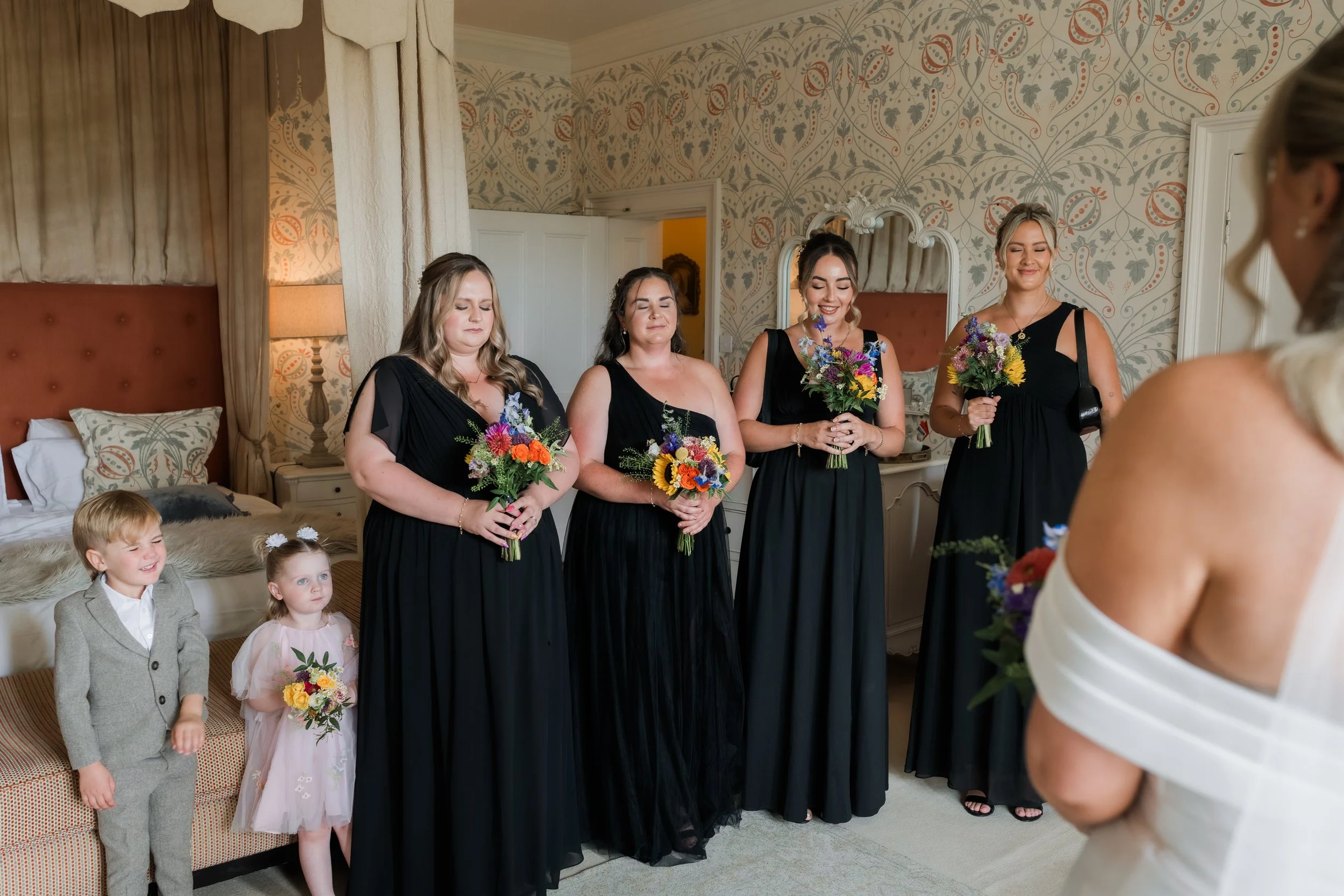 Group of four women in black dresses holding colorful bouquets, standing on a bed in a room with vintage wallpaper and a mirror. Two small children, a boy in a gray suit and a girl in a pink dress, stand nearby, smiling. A bride with blonde hair, wea