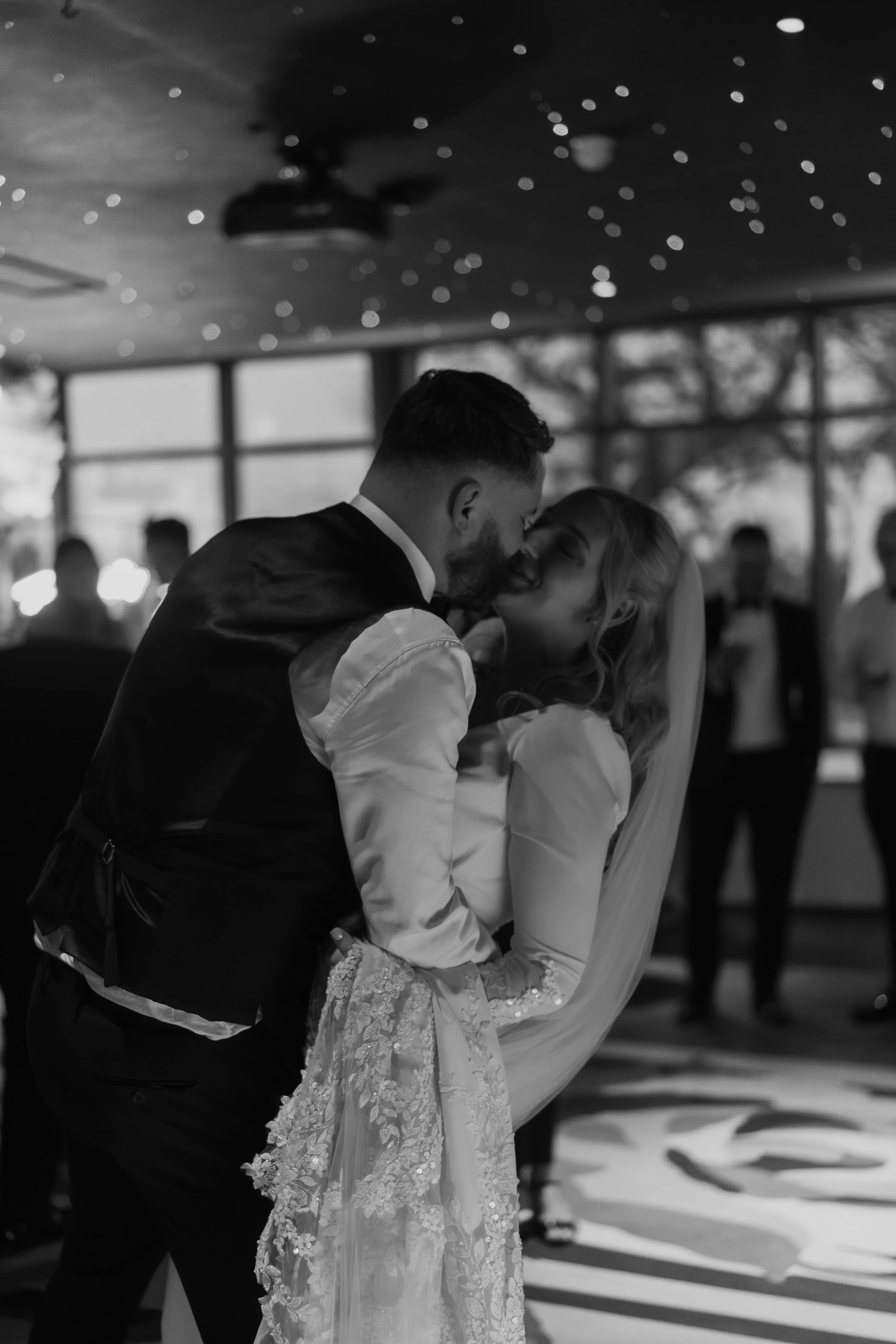 A black and white photo of a newlywed couple sharing a kiss while dancing at their wedding reception, with guests in the background and string lights on the ceiling.