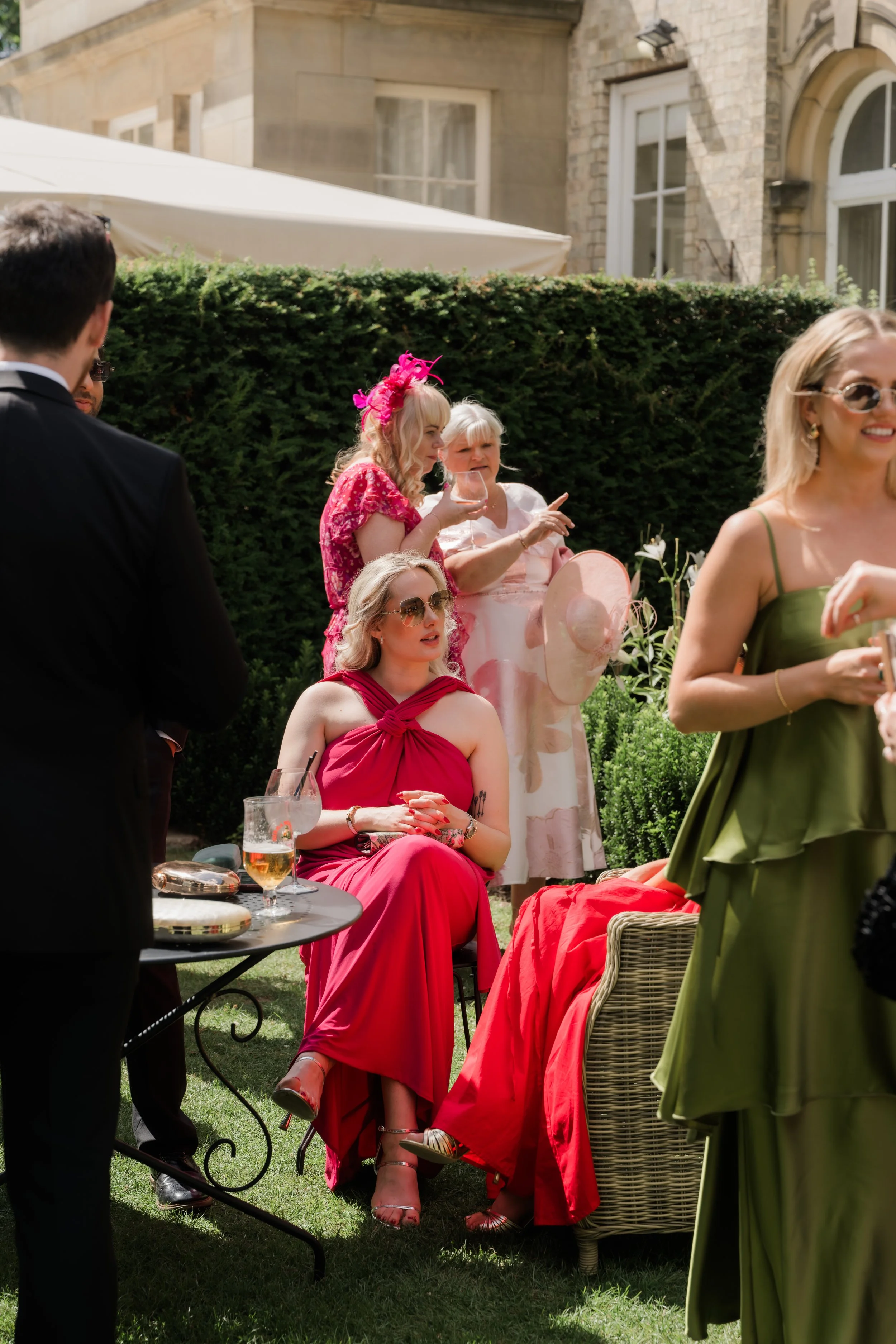 People socializing outdoors during a garden party, with some seated and others standing, enjoying drinks and conversation, in front of a hedge and a stone building.