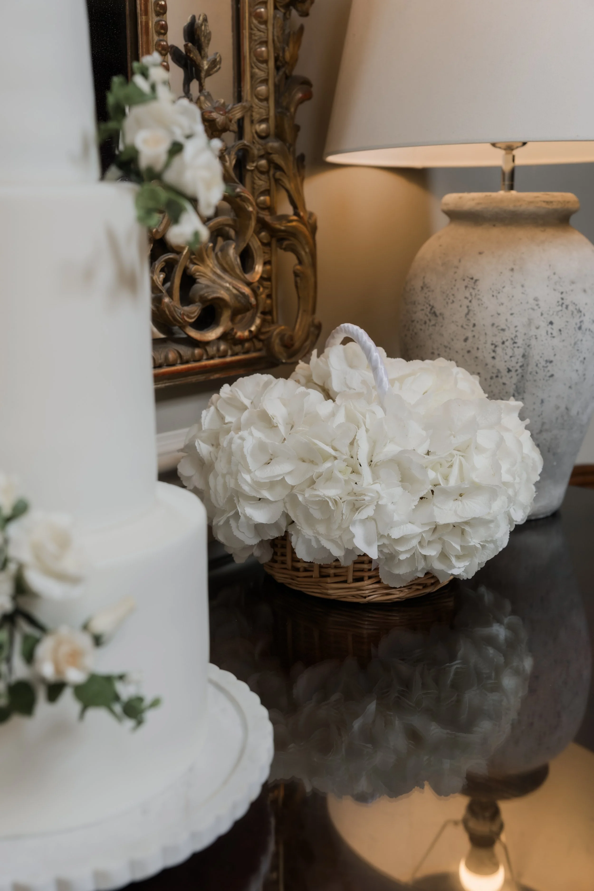 Close-up view of a decorated table with a white wedding cake, a wicker basket filled with white hydrangeas, a decorative ornate gold frame, and a large beige ceramic table lamp with a white shade.