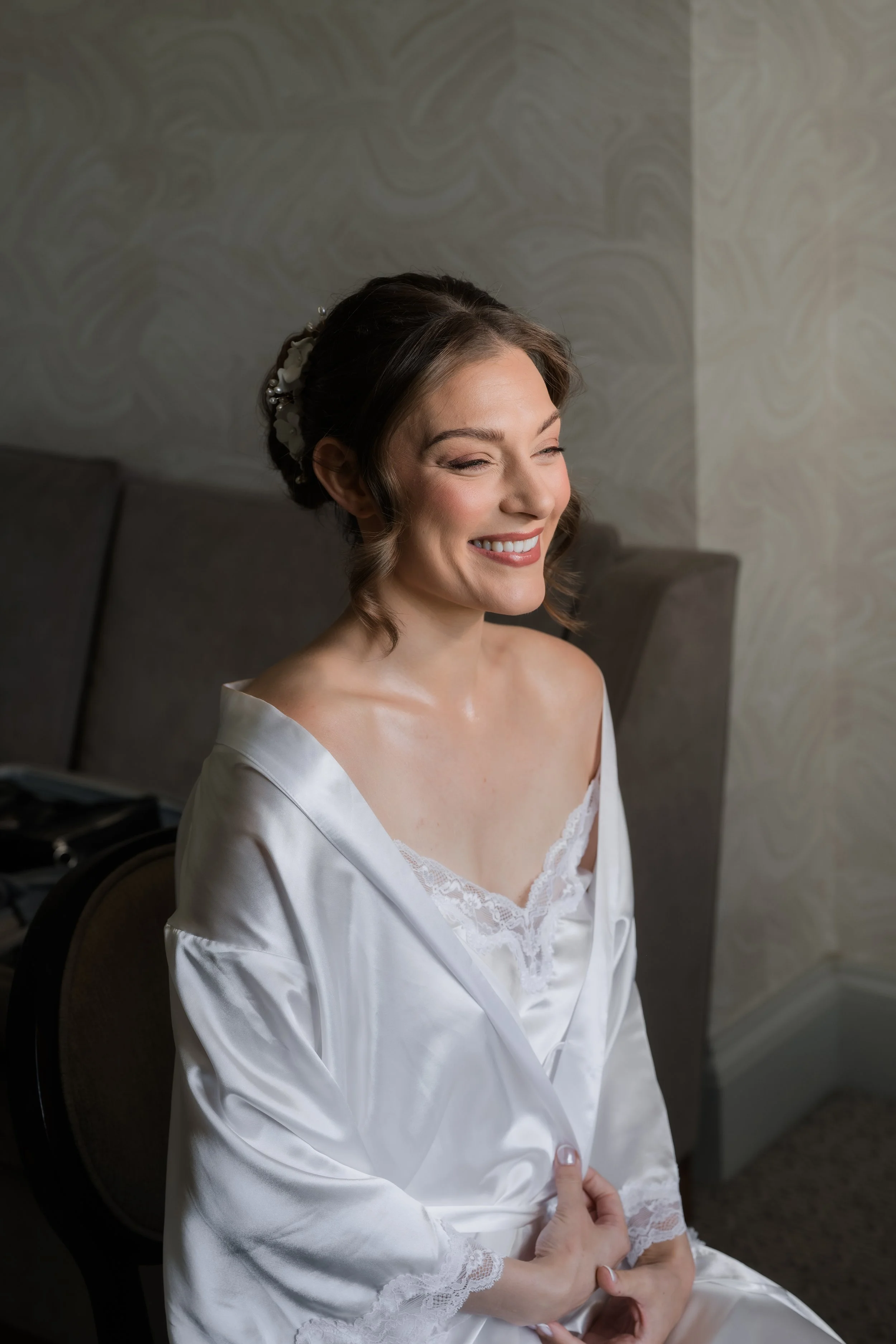A smiling woman in a satin robe with lace trim, sitting with her eyes closed and arms crossed, in a room with patterned wallpaper.