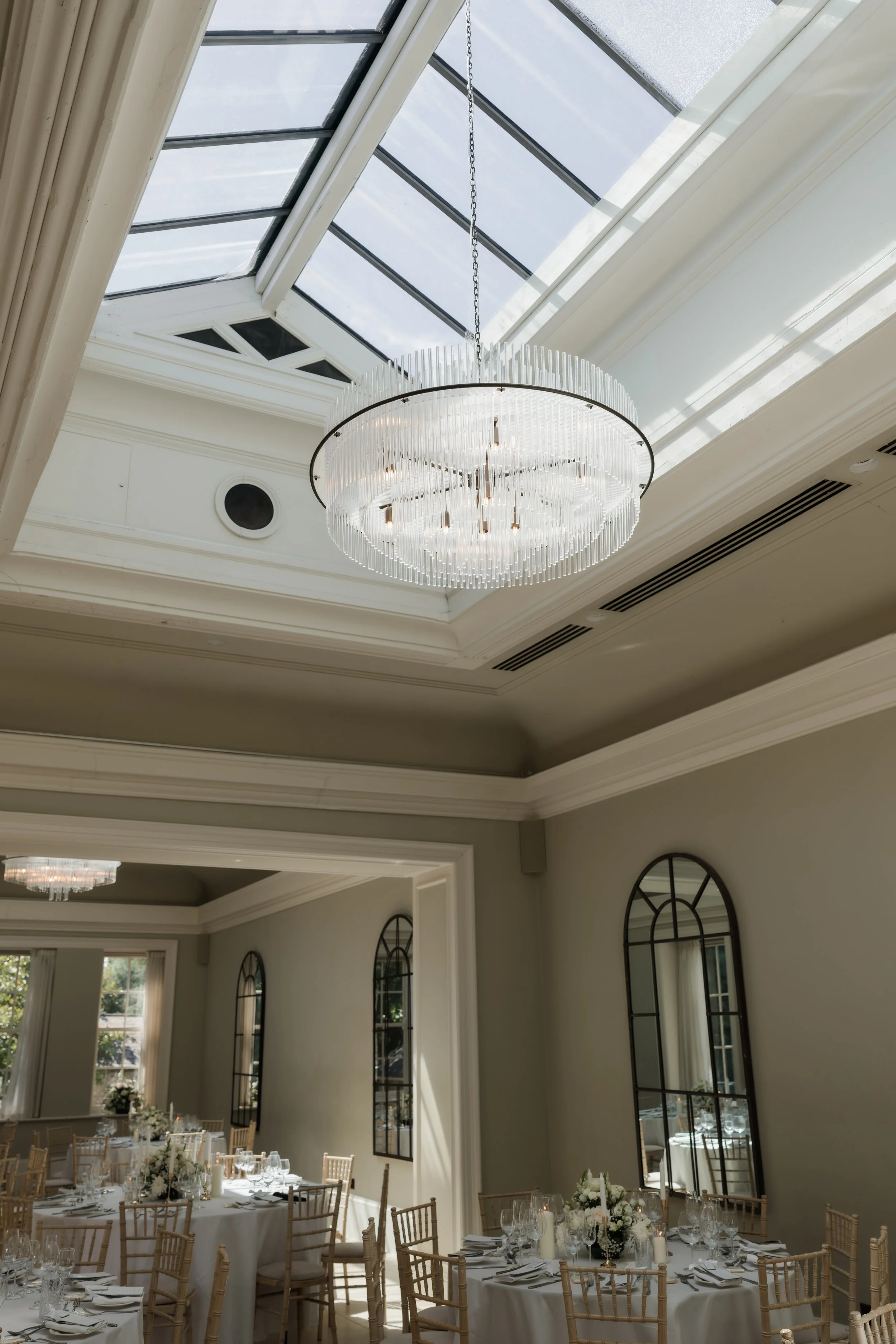 A formal event space with a large glass ceiling and a modern chandelier hanging from the ceiling. The room has elegant round tables set with white tablecloths, candles, and floral centerpieces, surrounded by wooden chairs. Mirrored windows line the w