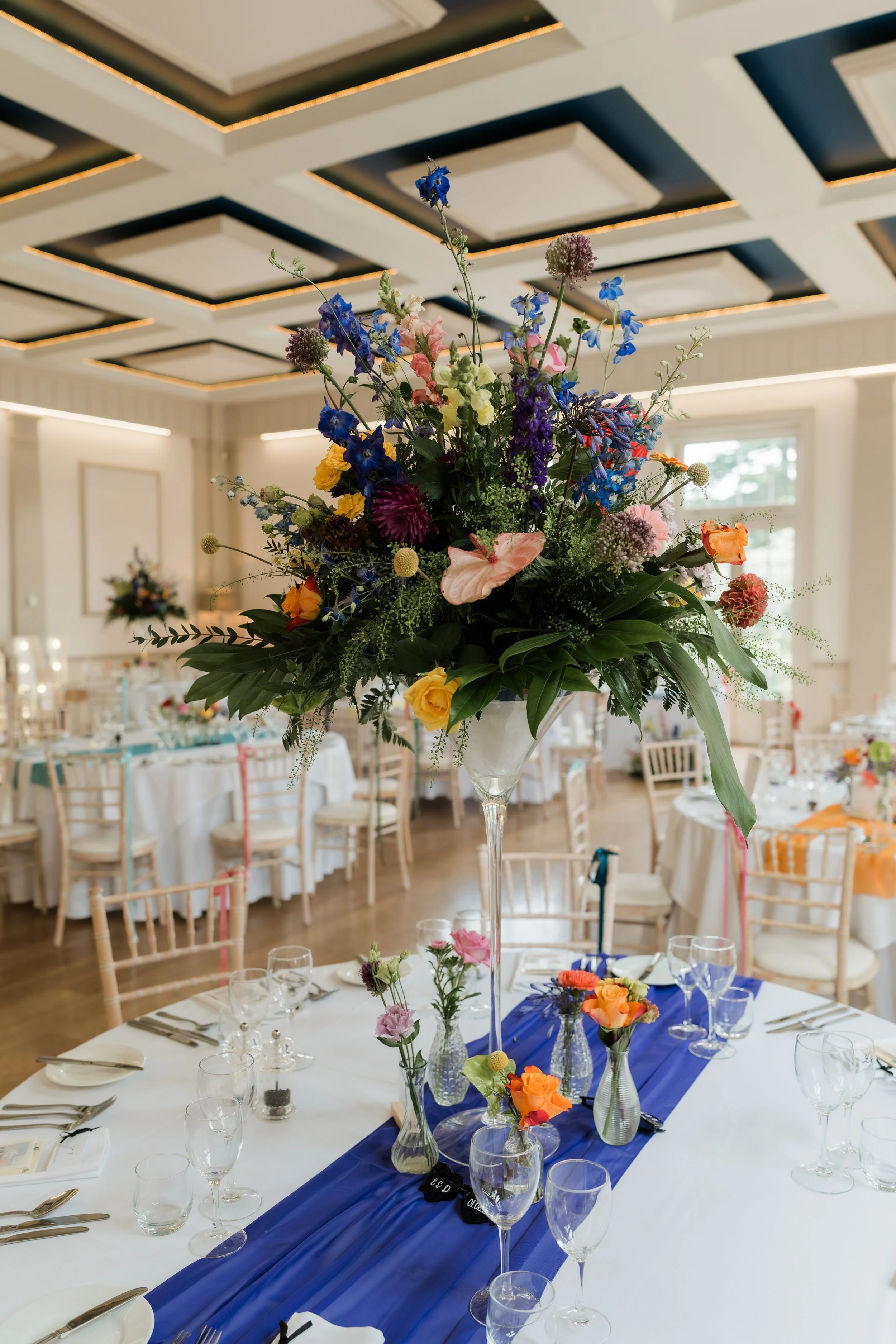 Large floral centerpiece with mixed colorful flowers on a dining table in a decorated event hall.