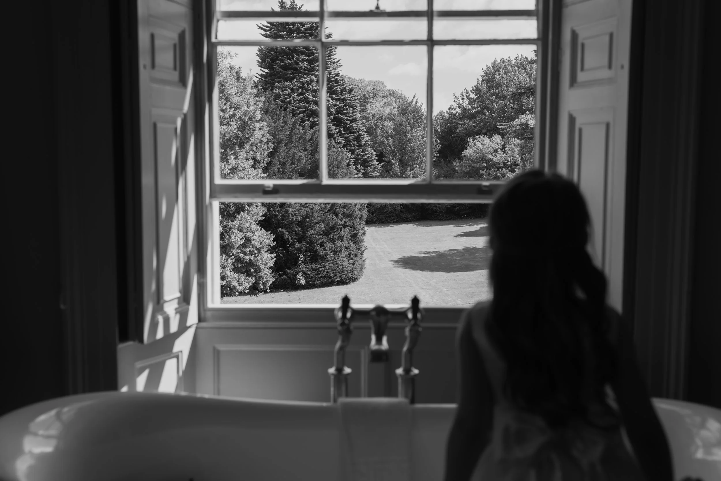 A black-and-white photo shows a girl looking out a large, multi-pane window onto a landscape of trees and a grassy area, with puzzle pieces on the ledge in the foreground.