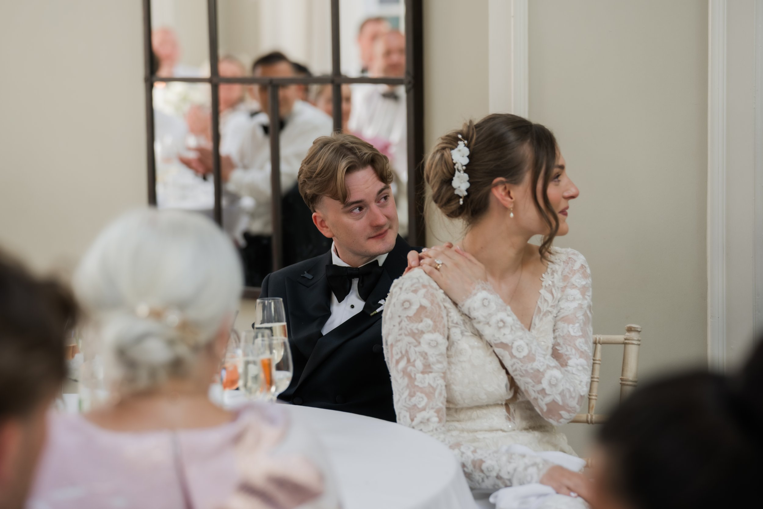 A bride and groom sit at a table during their wedding reception. The groom is wearing a black tuxedo and the bride is in a white lace wedding dress with floral hair accessories. There are guests and a mirror in the background.