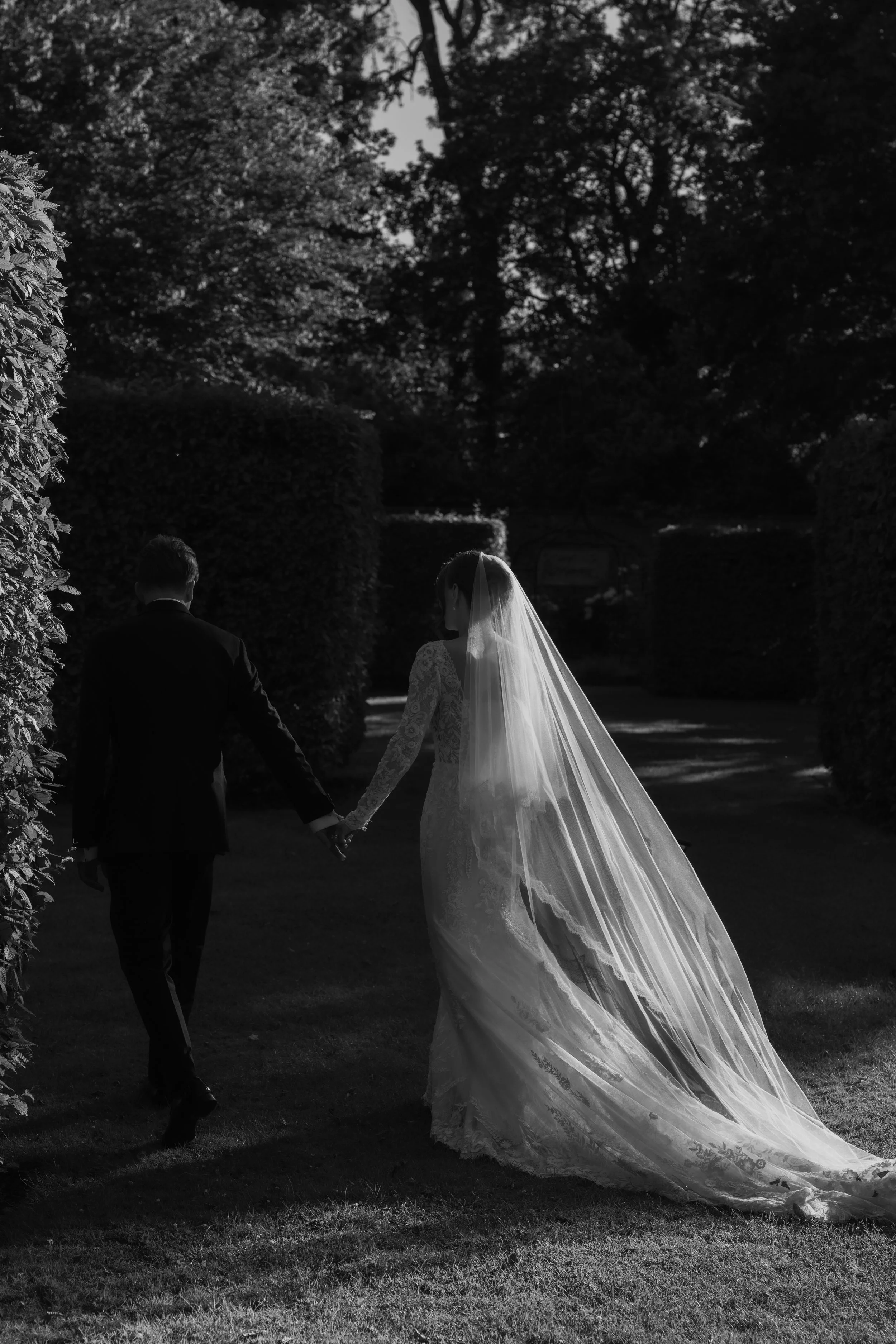 Black and white photo of a bride and groom holding hands, walking outdoors in a garden or park, with trees and bushes in the background.