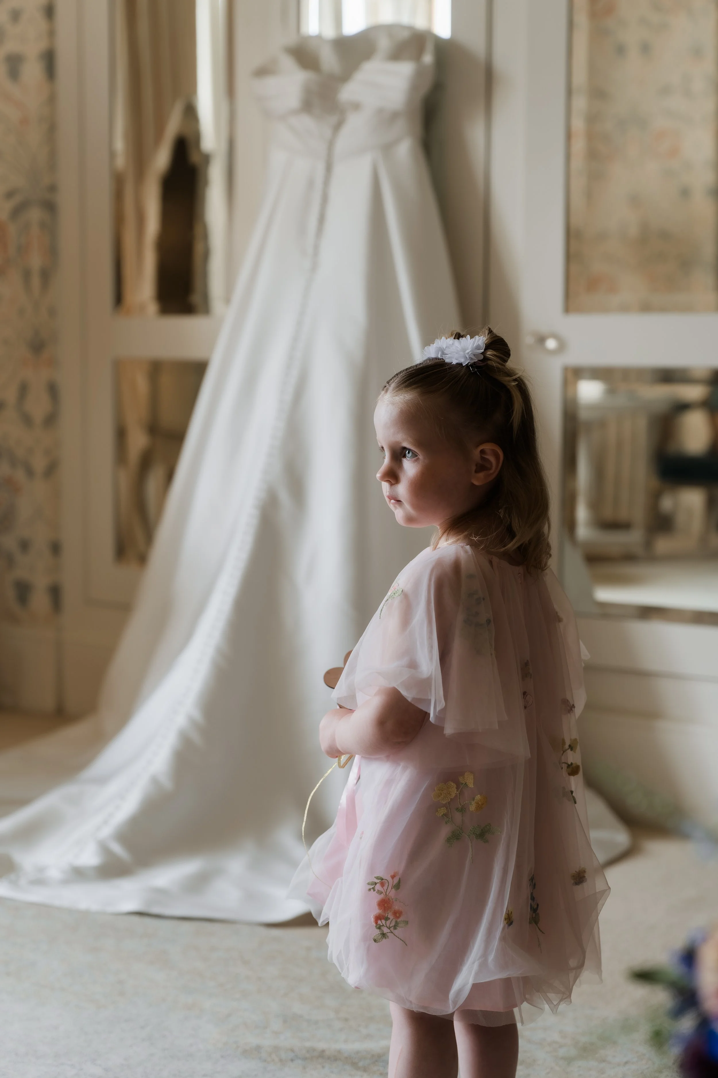 Young girl with a floral dress and top knot hair style looking at a white wedding dress on a mannequin in a well-lit room.