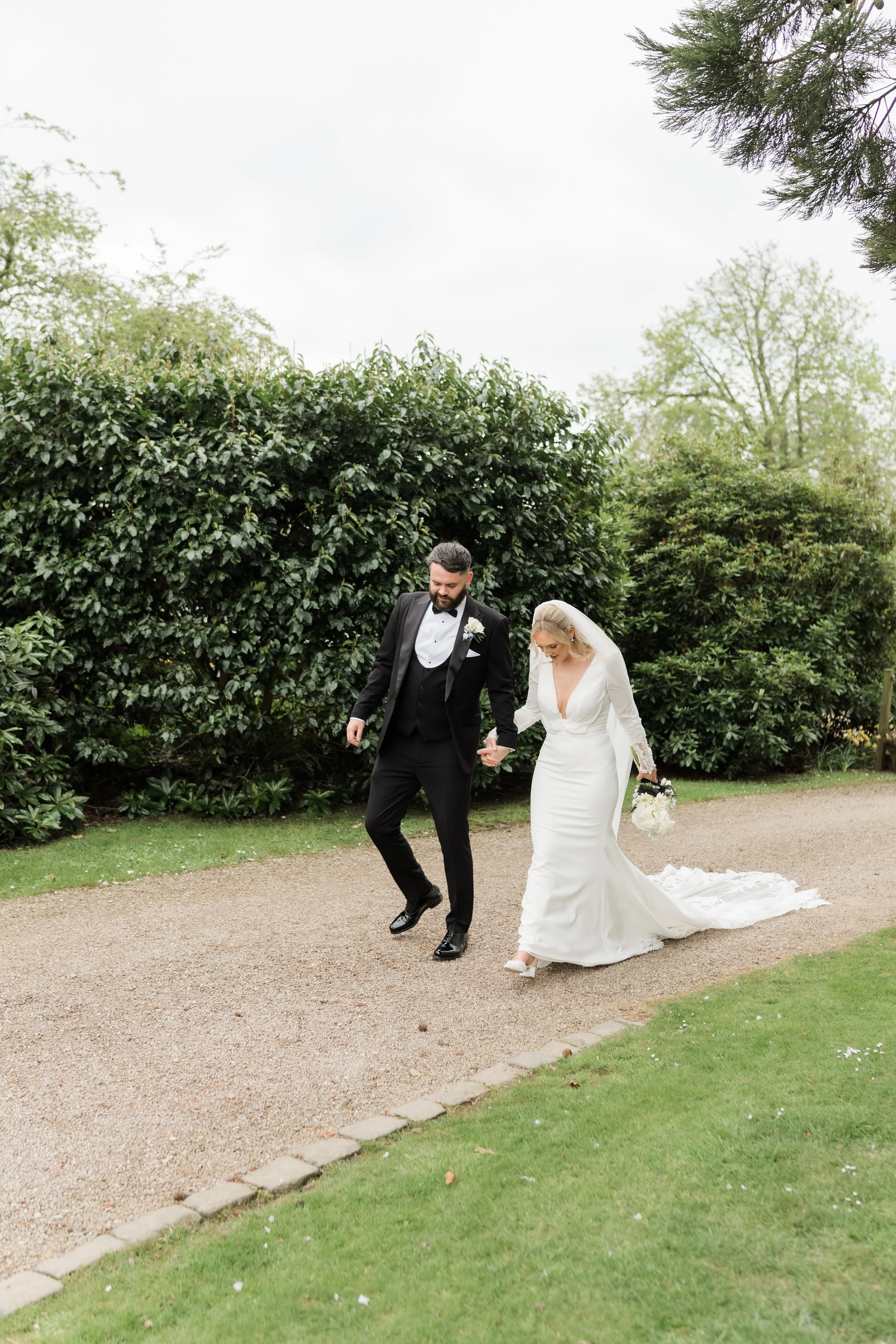 Bride and groom walking together outdoors on a wedding day, holding hands, with lush greenery in the background.