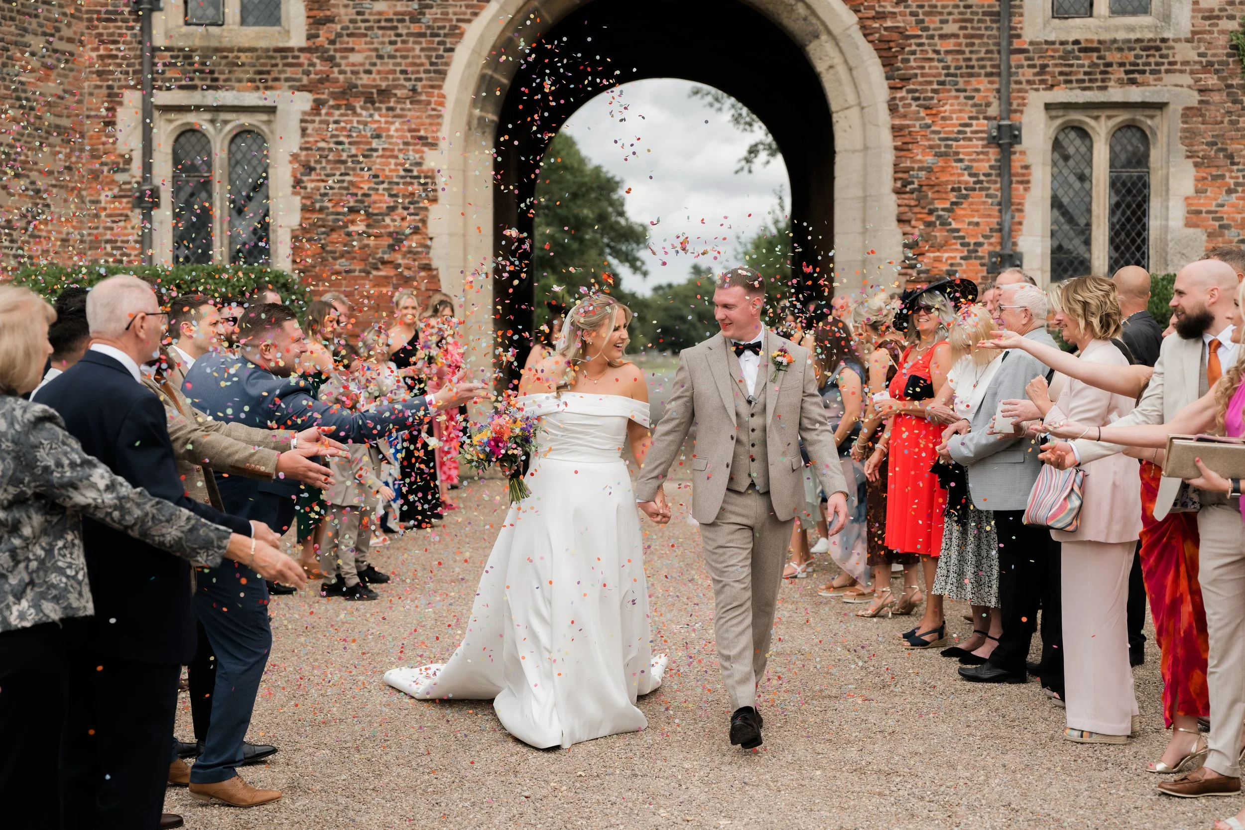 A wedding celebration with a bride and groom walking hand-in-hand through a colorful confetti shower, surrounded by guests outside a brick building.