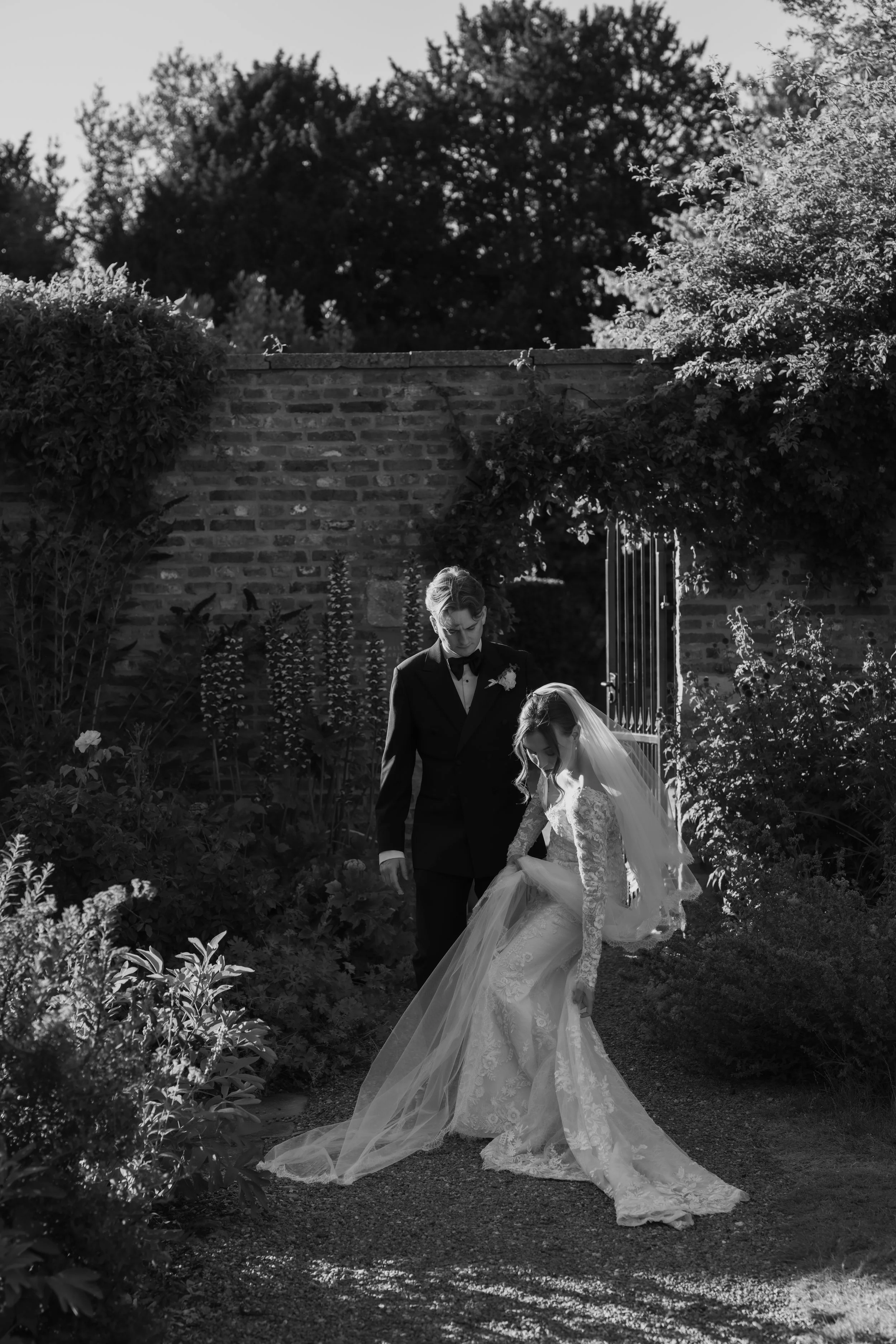 A bride and groom walking together in a garden, with the bride holding up her wedding dress and the groom looking down at her, surrounded by lush plants and trees, with a brick wall and gate in the background.