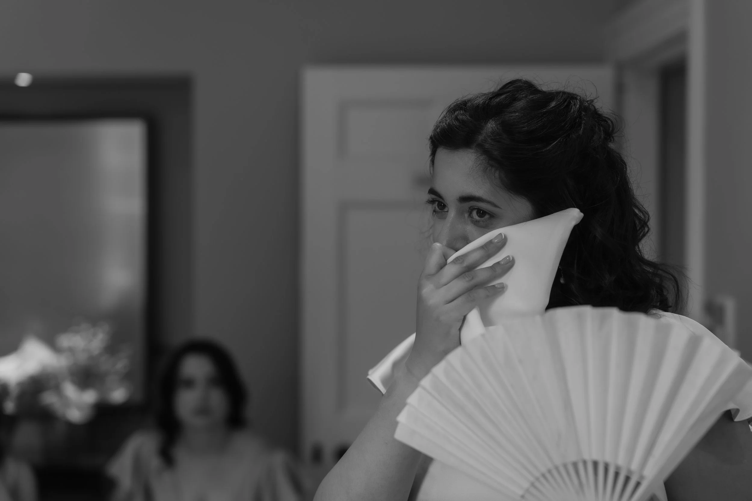 A woman with dark curly hair holding a tissue to her nose, near a folding fan, with another woman in the background looking away in a room.