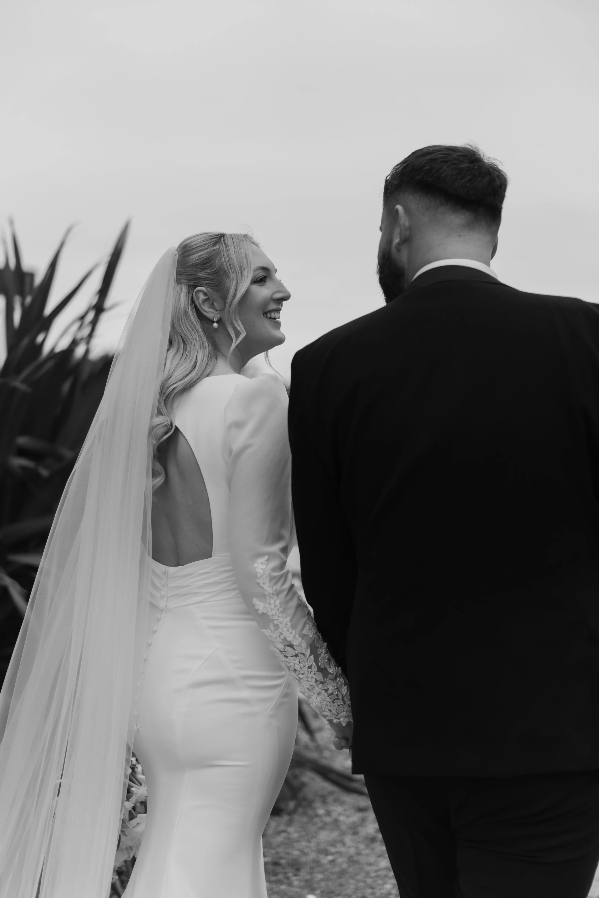 A black and white photograph of a bride and groom holding hands outdoors, facing each other, with a cloudy sky in the background.