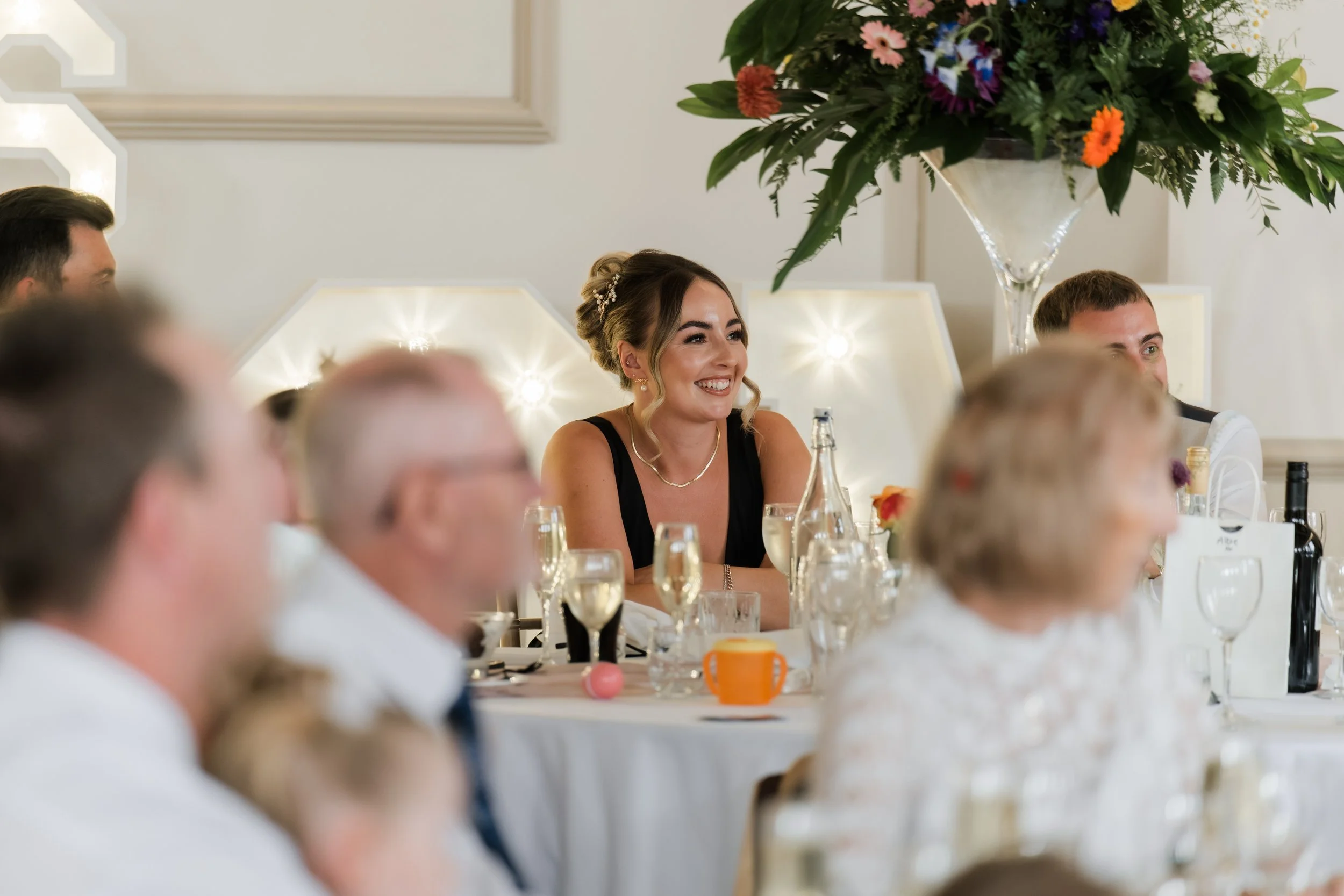 A woman in a black dress smiling at a formal event, surrounded by other guests and a large floral arrangement.