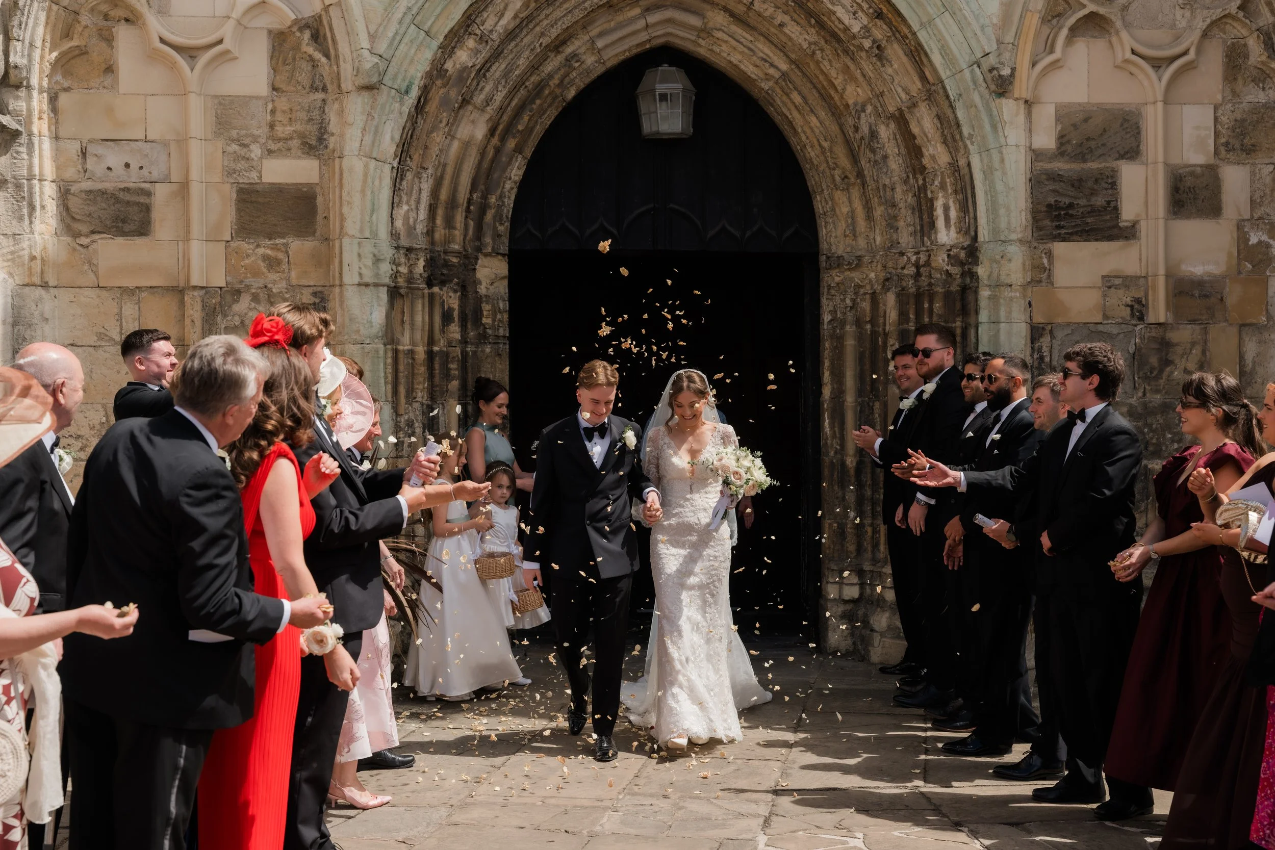 A wedding ceremony taking place outside a church with a stone facade. The bride and groom are walking out of the church, surrounded by guests throwing flower petals.