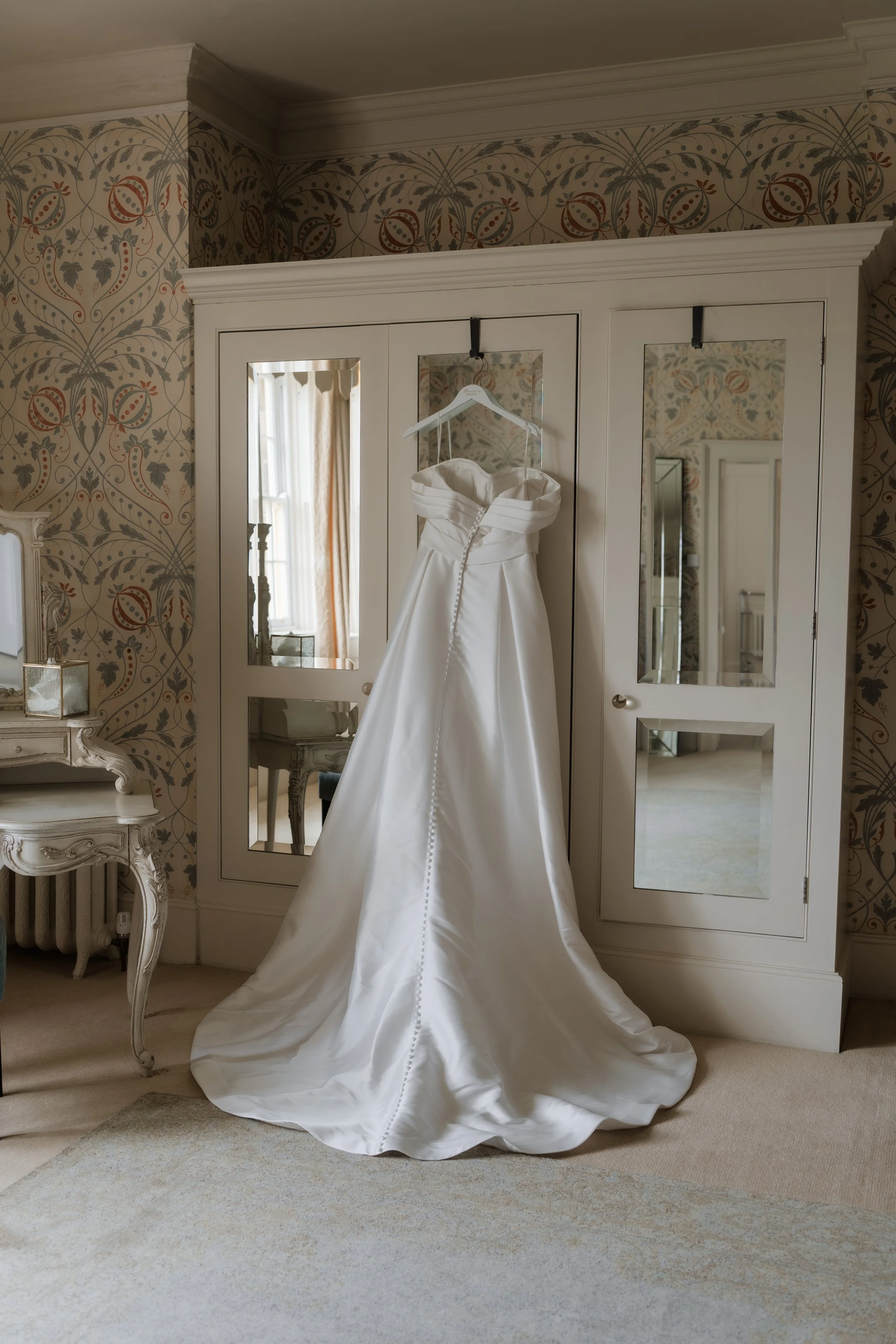 A white wedding dress hanging on a hanger inside a wardrobe in a room with floral wallpaper and a window with curtains.