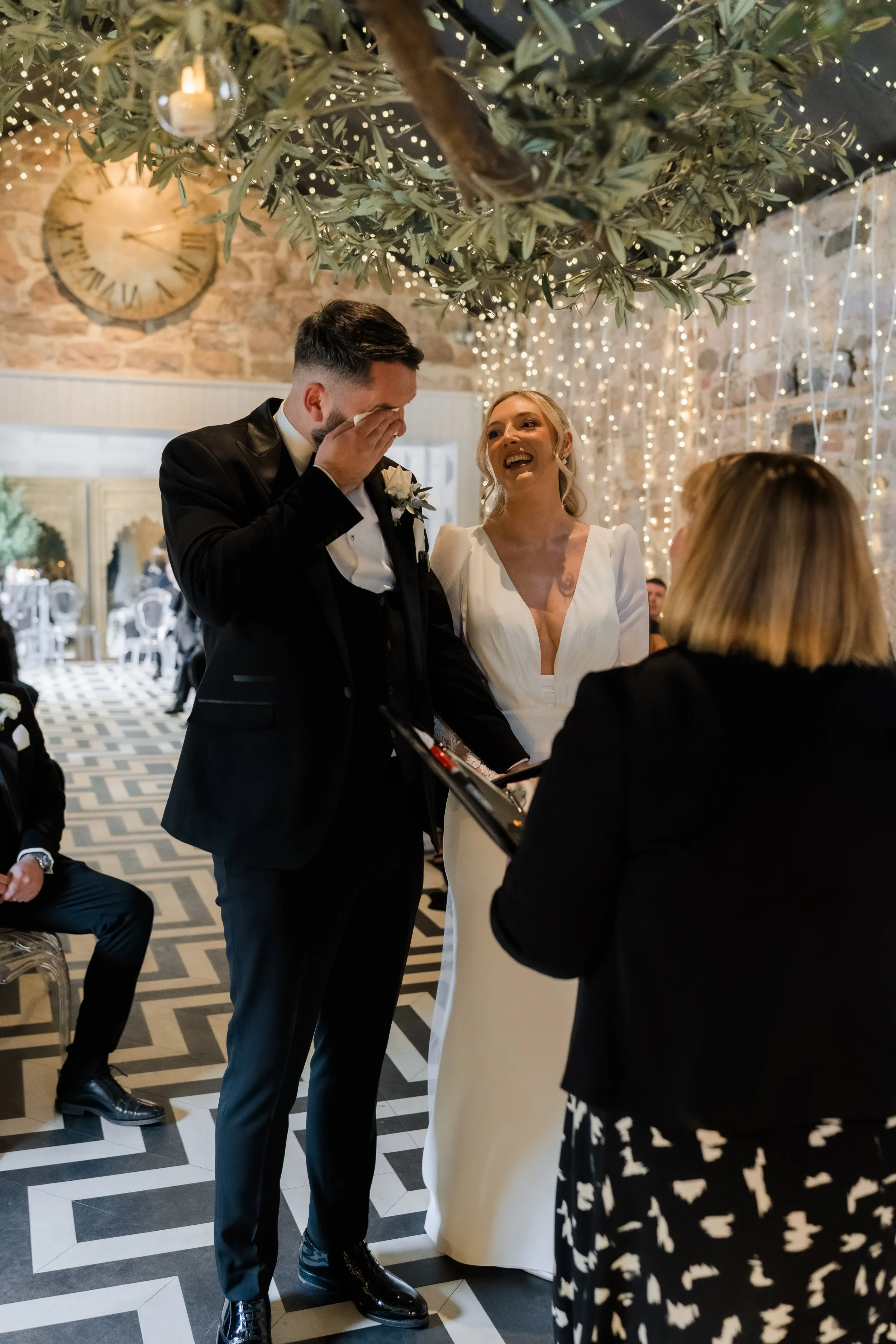 A wedding ceremony with a bride and groom smiling, standing under a hanging olive branch decorated with string lights, as an officiant reads from a folder. The groom is wiping away tears, and the bride is laughing. There are guests in the background 