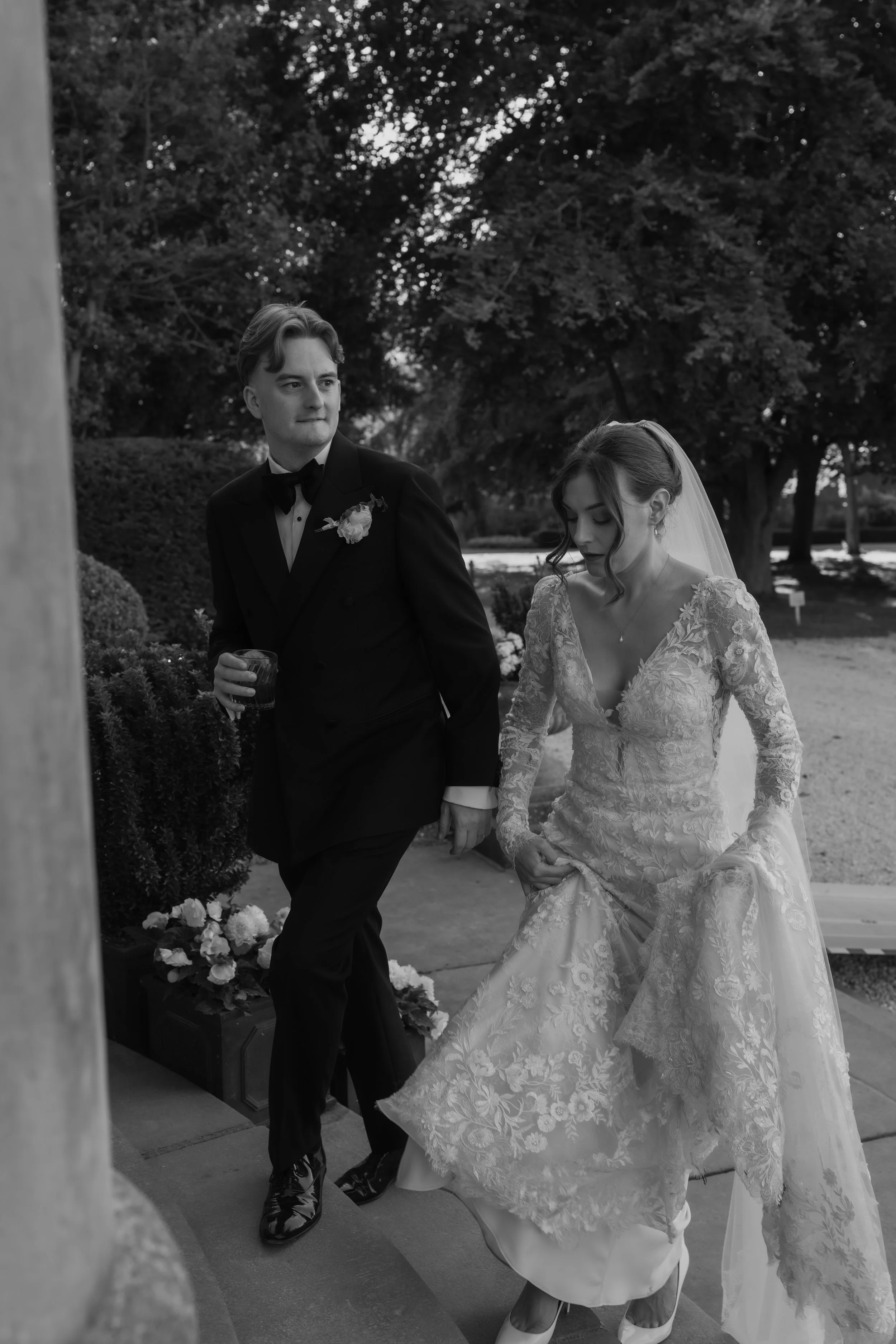 A black and white photo of a wedding scene showing a groom in a tuxedo holding a glass, standing beside a bride in a lace wedding gown with a veil, outdoors with trees and bushes in the background.