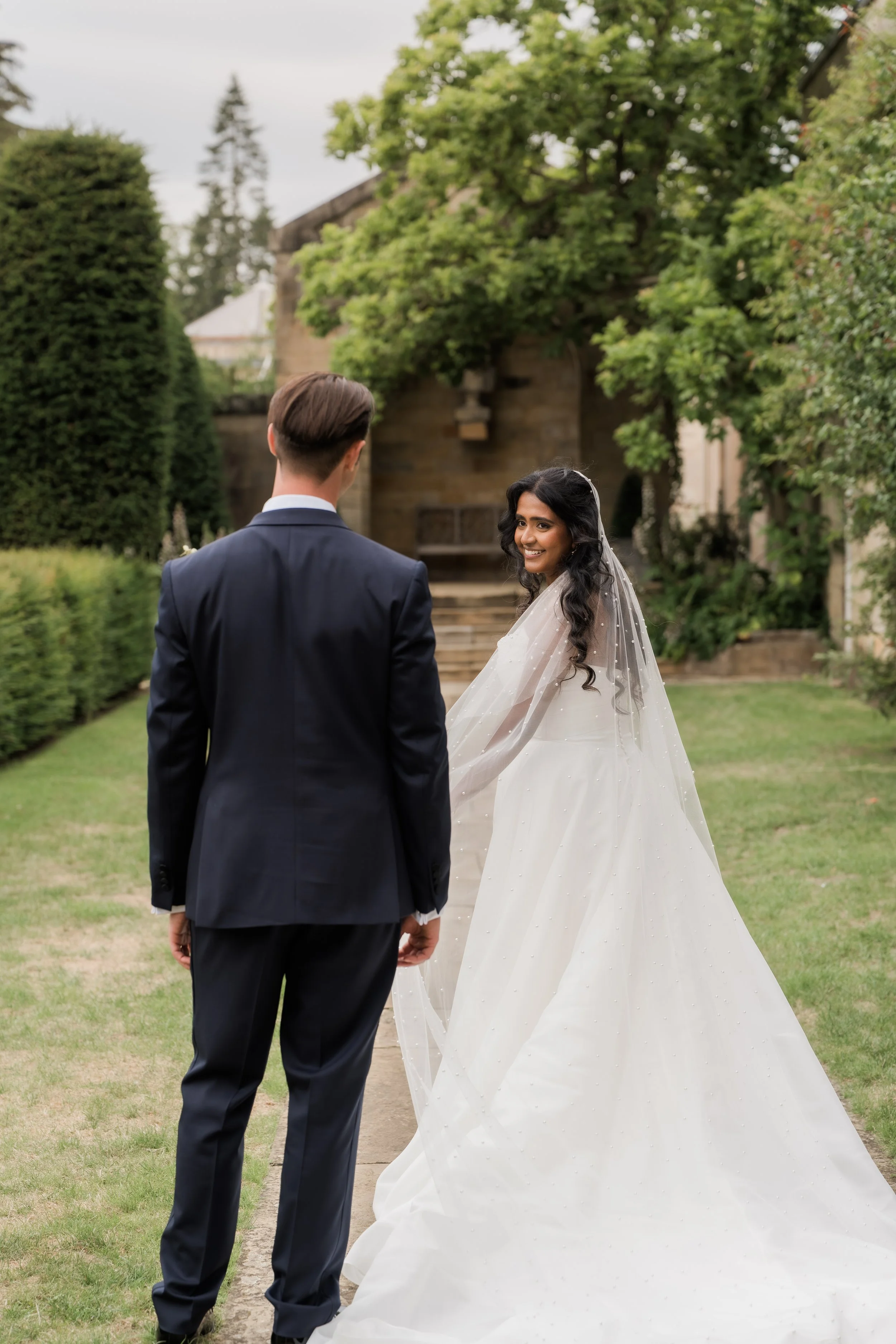 A bride and groom standing in a garden, holding hands, with the bride smiling at the camera and wearing a white wedding gown with a veil. The groom is dressed in a dark suit, with trees and a building in the background.