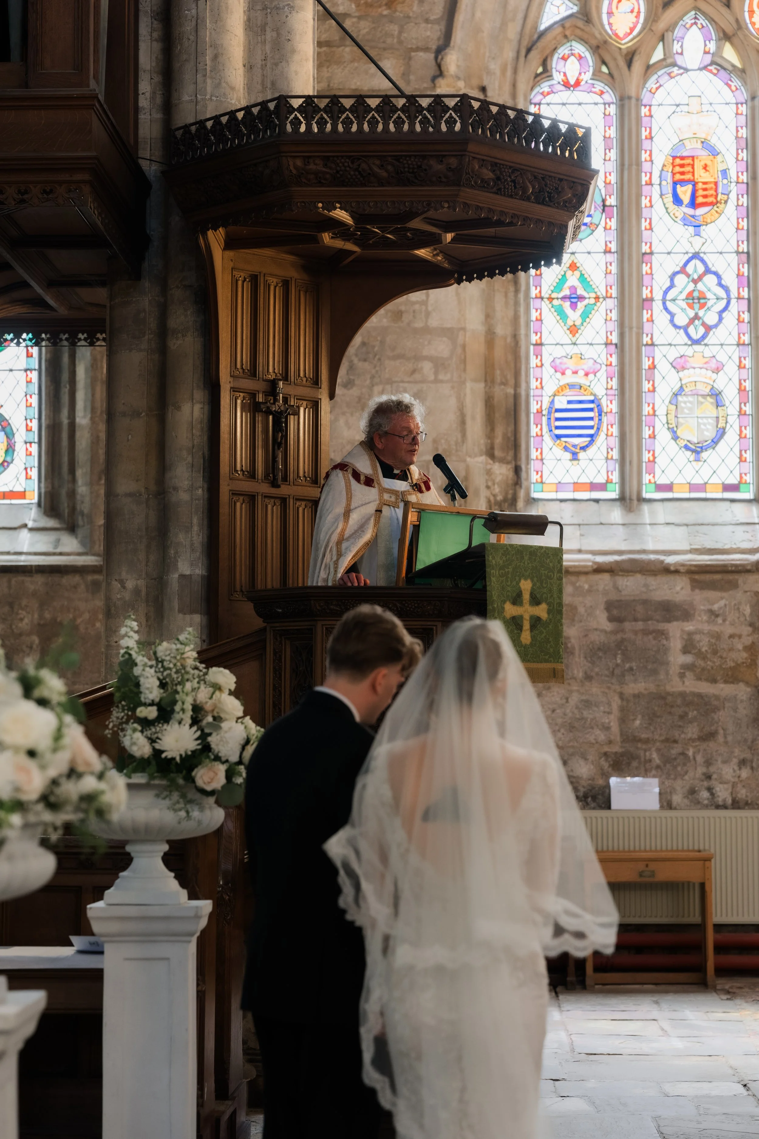 A wedding ceremony inside a church with stained glass windows, a priest in white robes, and a bride and groom standing at the altar. Floral arrangements decorate the space.