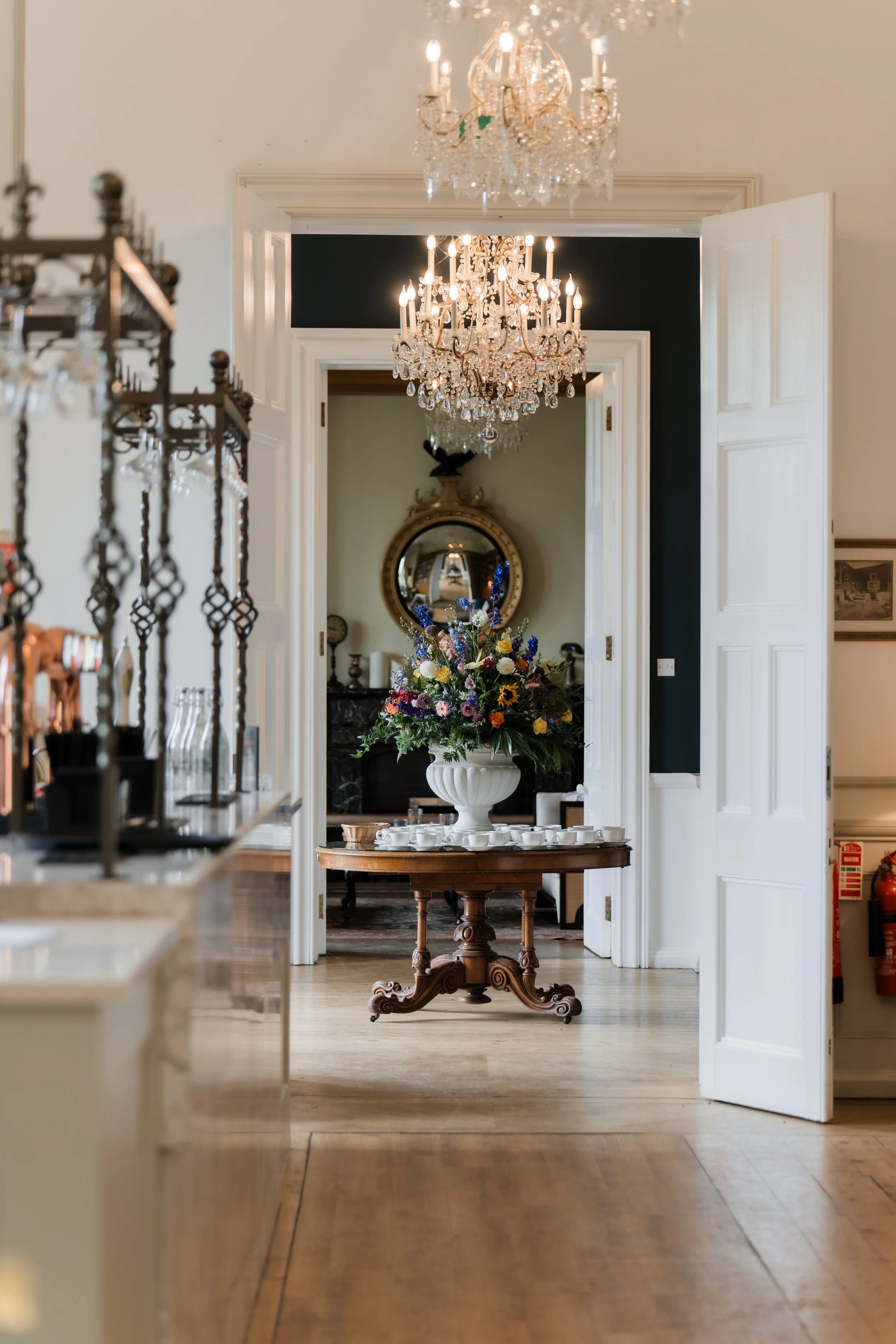 Elegant room with chandeliers, a large floral arrangement on a wooden table, and a mirror reflecting parts of the room.