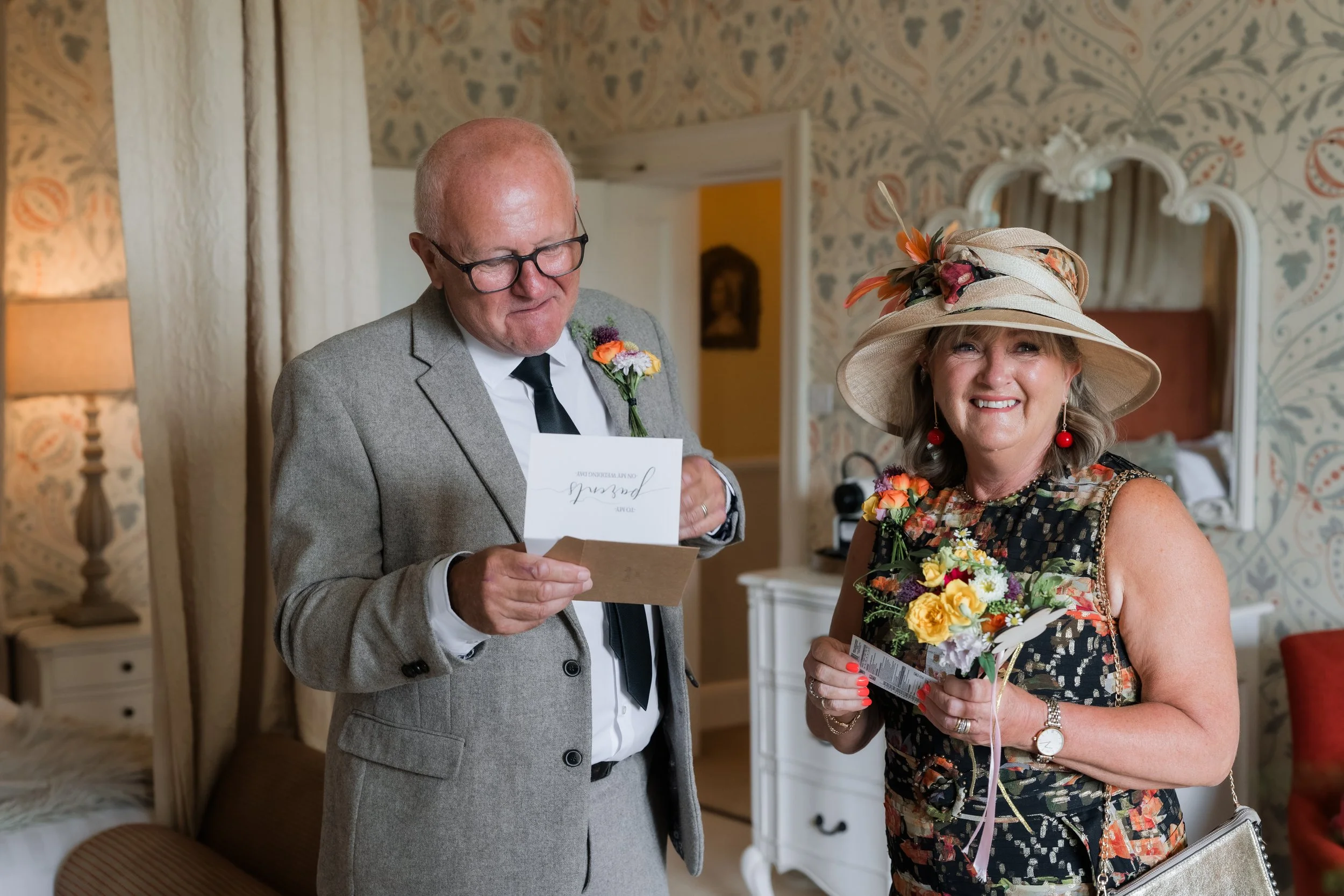 A smiling woman in a floral dress and large hat holding a bouquet of flowers, standing next to a man in a gray suit reading a card in a decorated room.