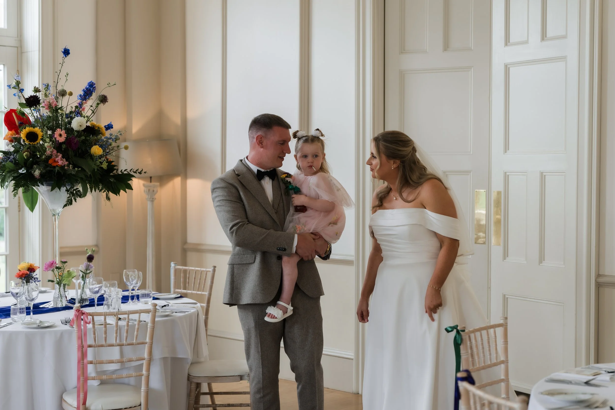 A couple at their wedding standing in a decorated room, the groom holding a young girl, with a woman in a white dress looking at them.