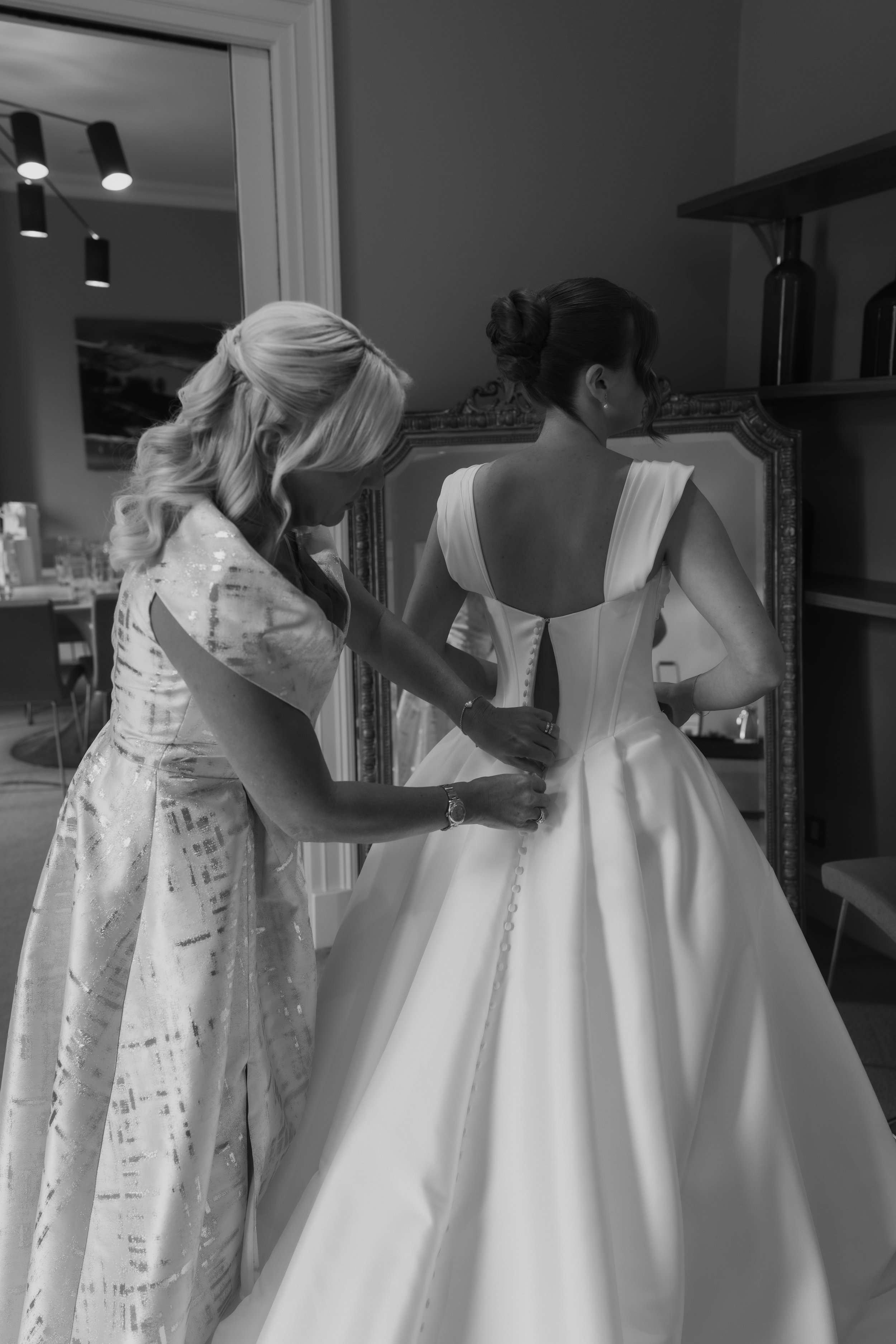 A woman assisting a bride with her wedding dress in a dressing room, both looking at the mirror.