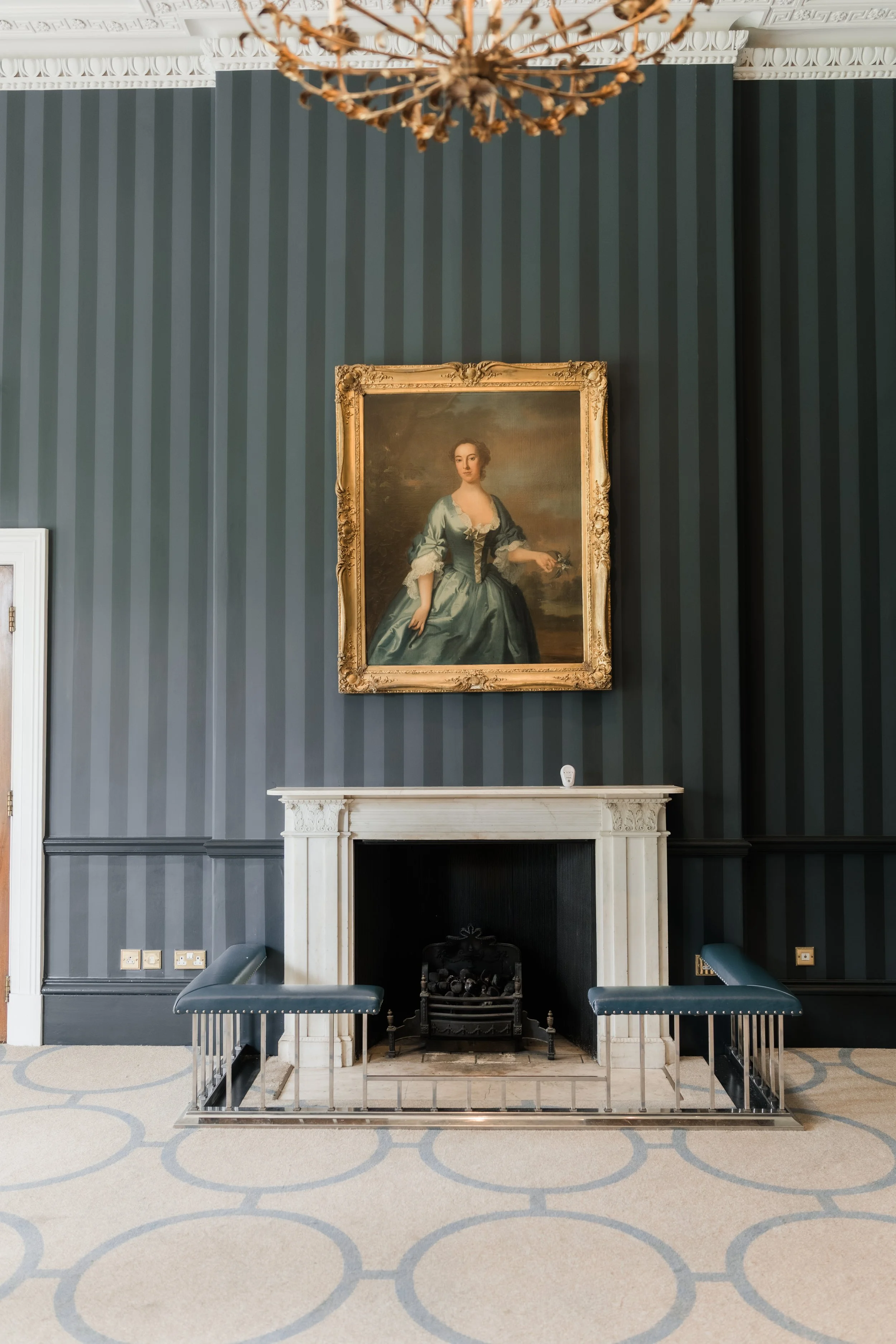 An elegant room with dark striped wallpaper, a white fireplace, and a portrait of a woman in historical attire hanging above the mantel. Two modern benches are positioned on either side of the fireplace, and there is a ornate chandelier hanging from 