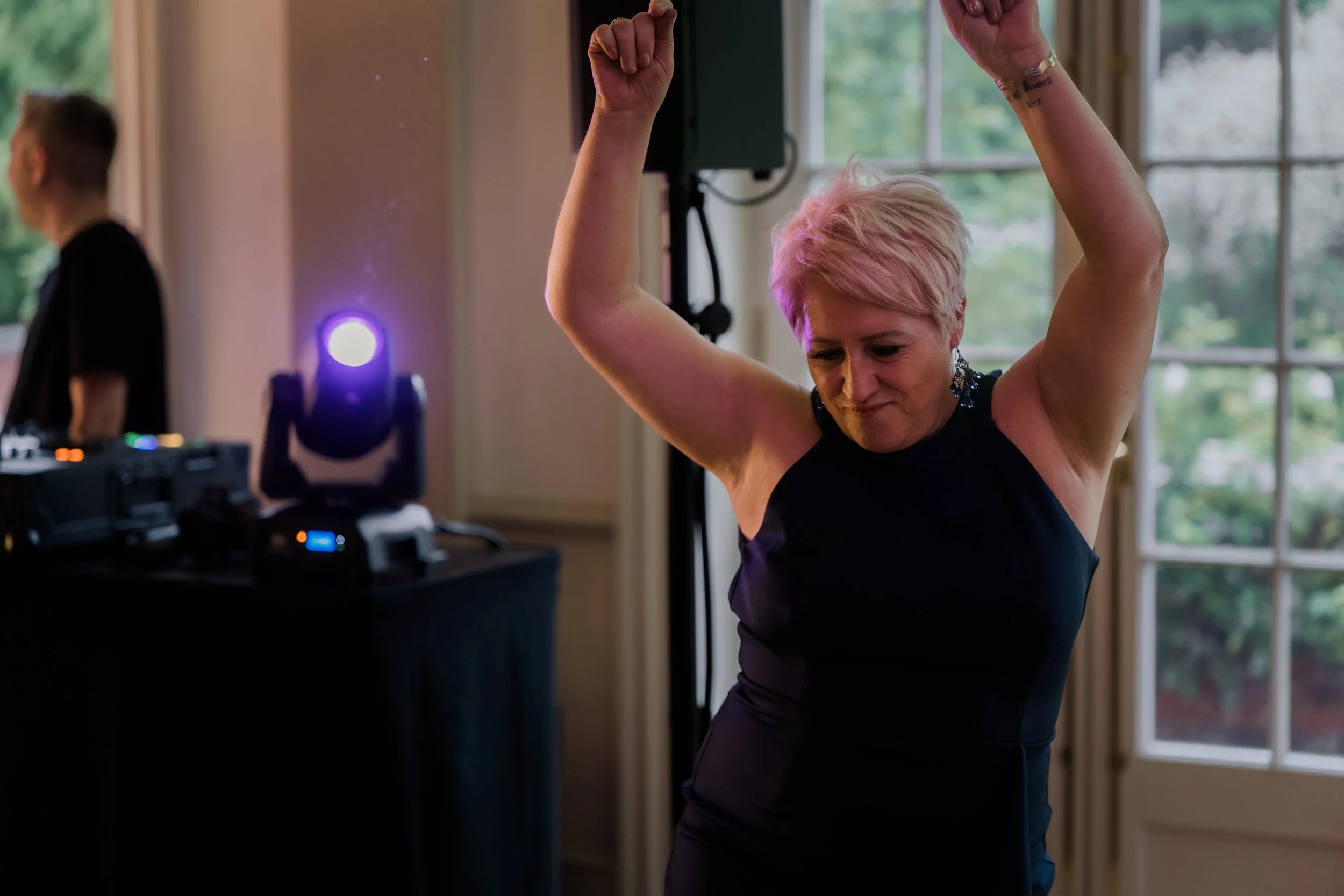 A woman with short pink hair dancing with her arms raised at an indoor event, with a DJ and audio equipment visible in the background.
