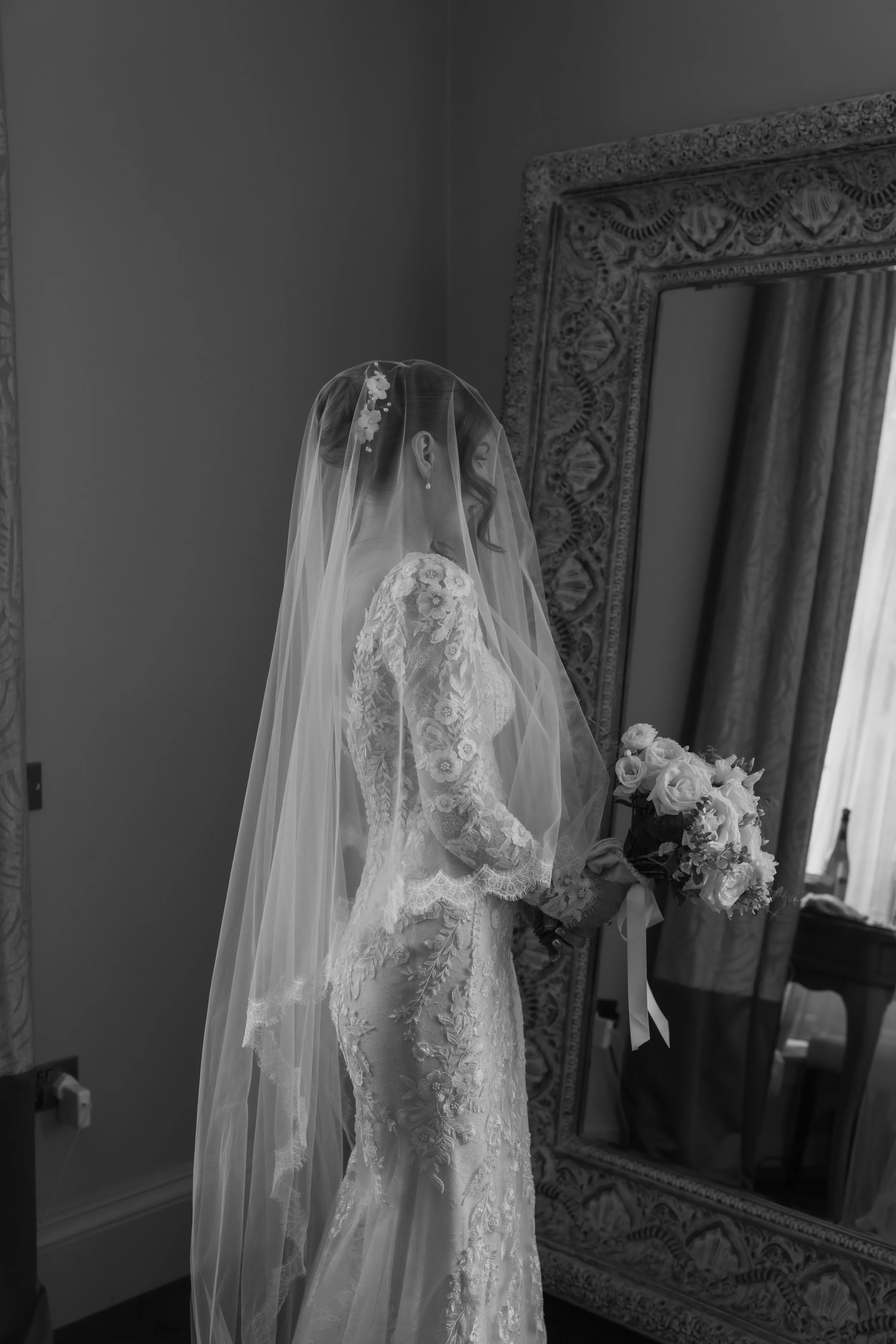 Black and white photo of a bride in lace wedding dress with veil holding a bouquet of white roses, standing in front of a large ornate mirror and curtains.