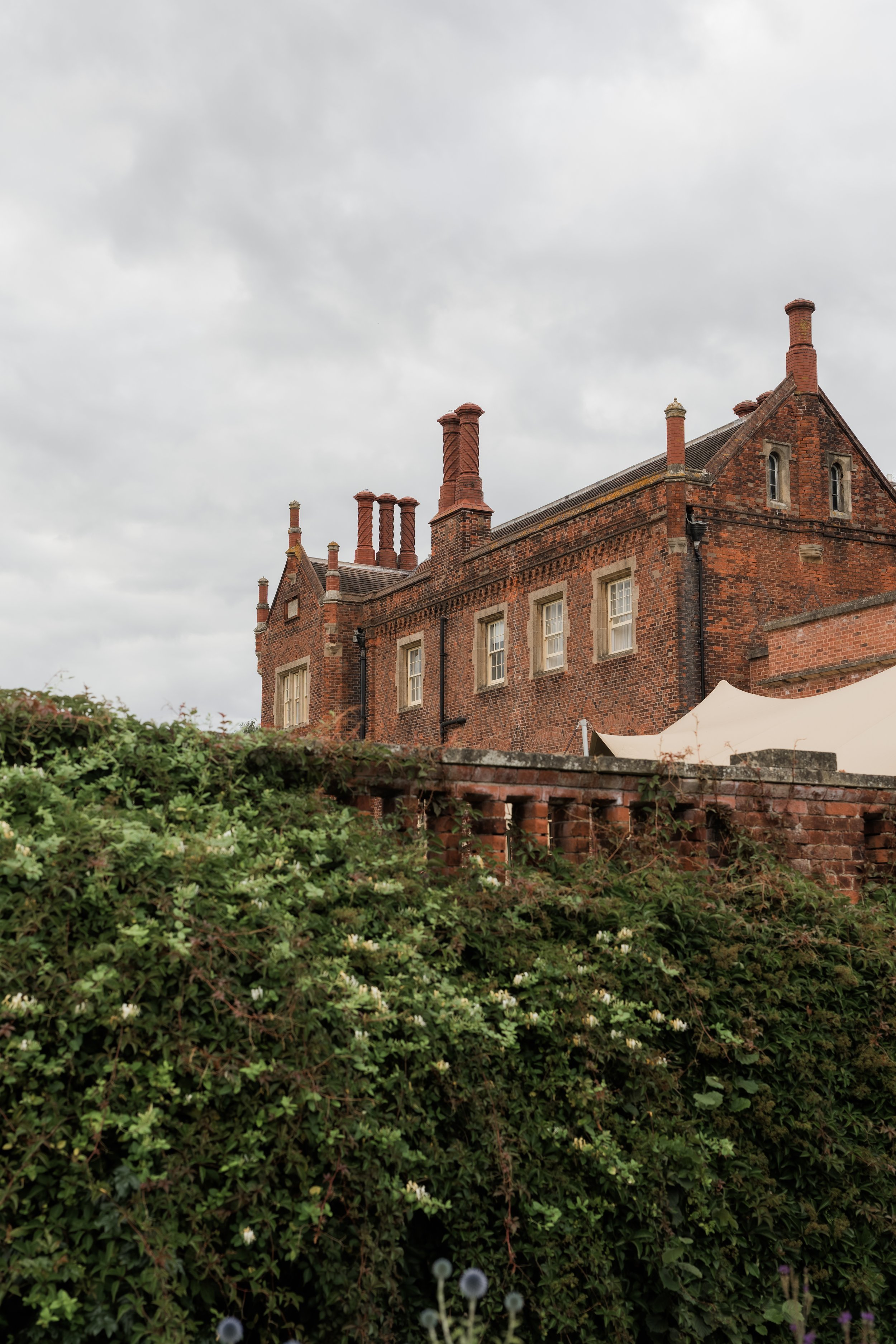 A large red brick house with multiple chimneys, small windows, and steeply pitched roof, under a cloudy sky, with a retaining wall and green foliage in the foreground.