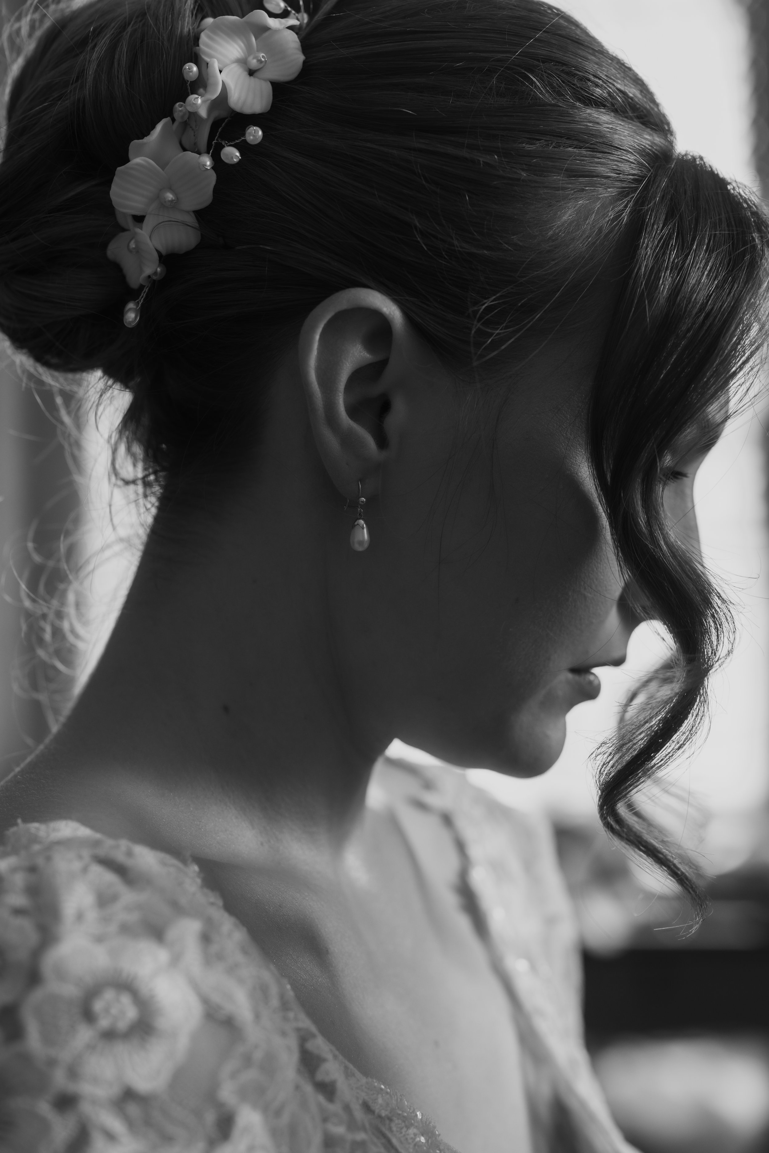 A close-up black and white photograph of a woman’s profile, showing her face, an earring, her hair with decorative flowers and beads, and a lace garment she is wearing.