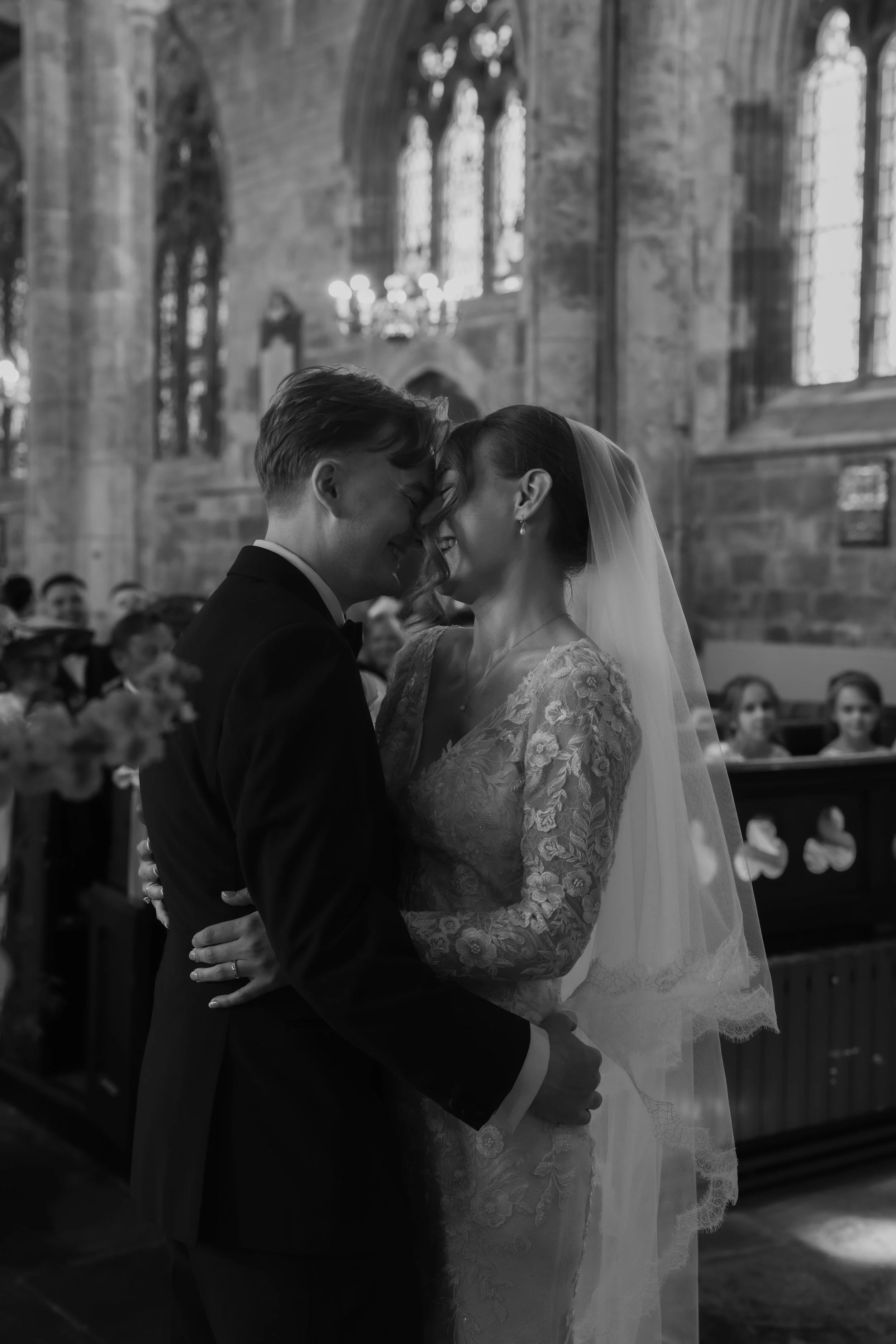 A black and white photo of a bride and groom sharing a kiss inside a church, with guests in the background.