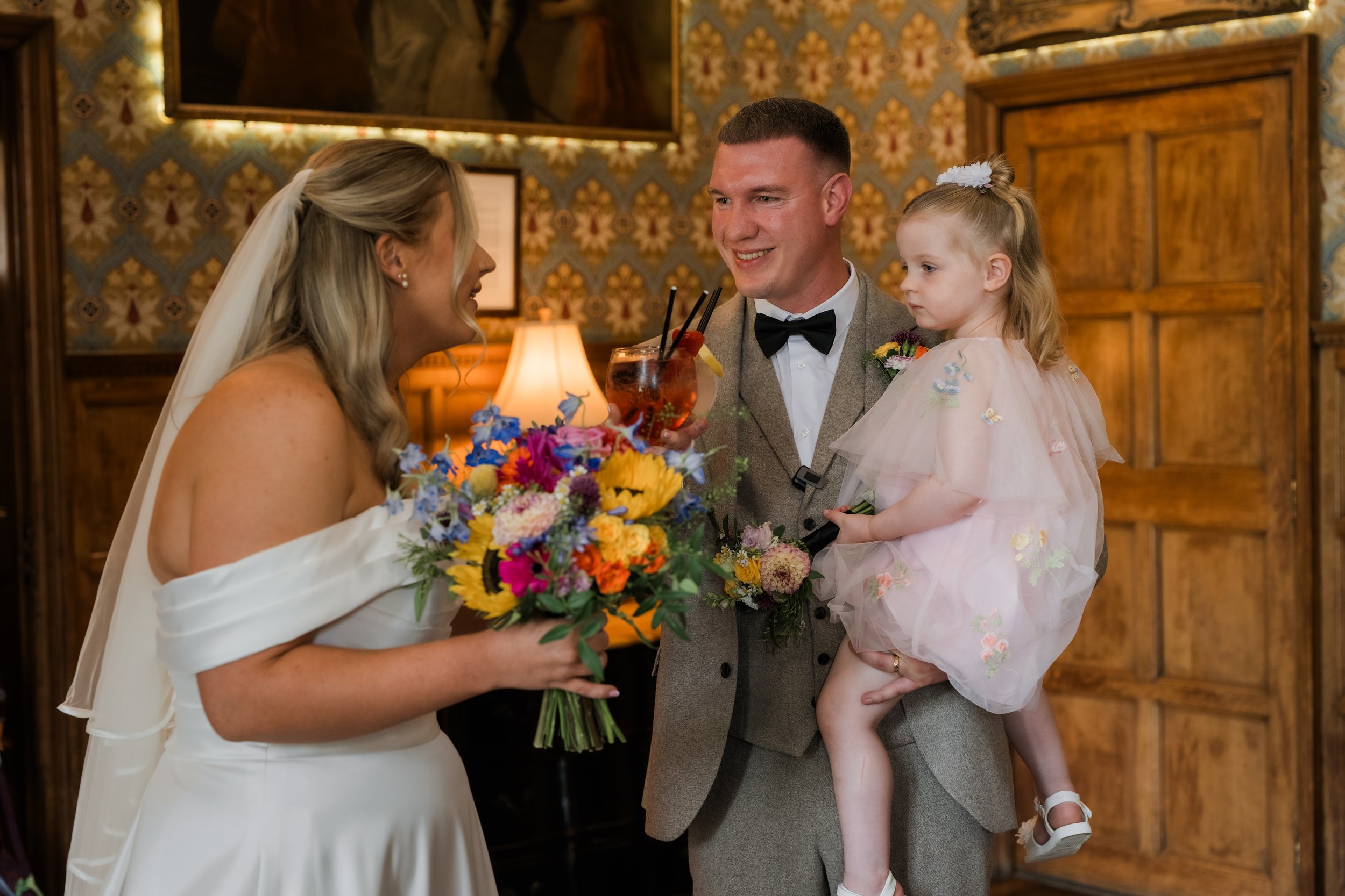 A bride, groom, and a young girl at a wedding reception. The bride is holding a colorful bouquet, the groom is holding a drink, and the girl is dressed in a pink dress with floral decorations. They are smiling and talking in a warmly decorated room.