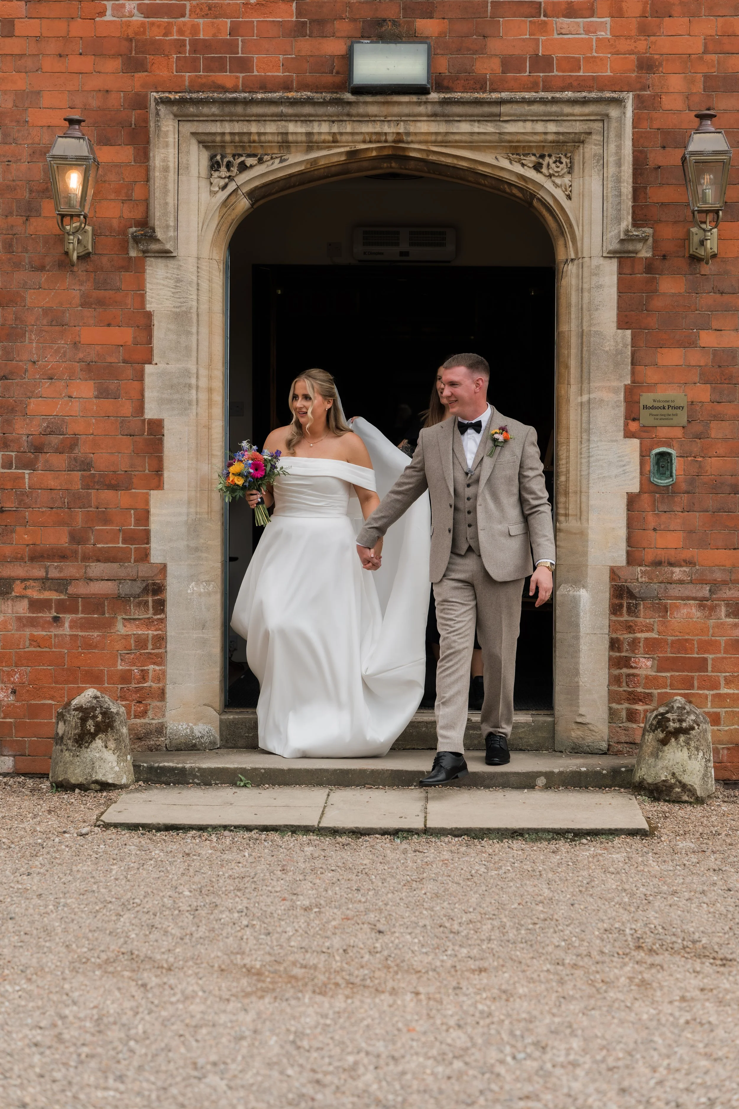 A bride and groom holding hands and smiling as they exit a red brick church. The bride is wearing a white off-shoulder wedding gown and holding a colorful bouquet, and the groom is dressed in a beige suit with a black bow tie and boutonniere.
