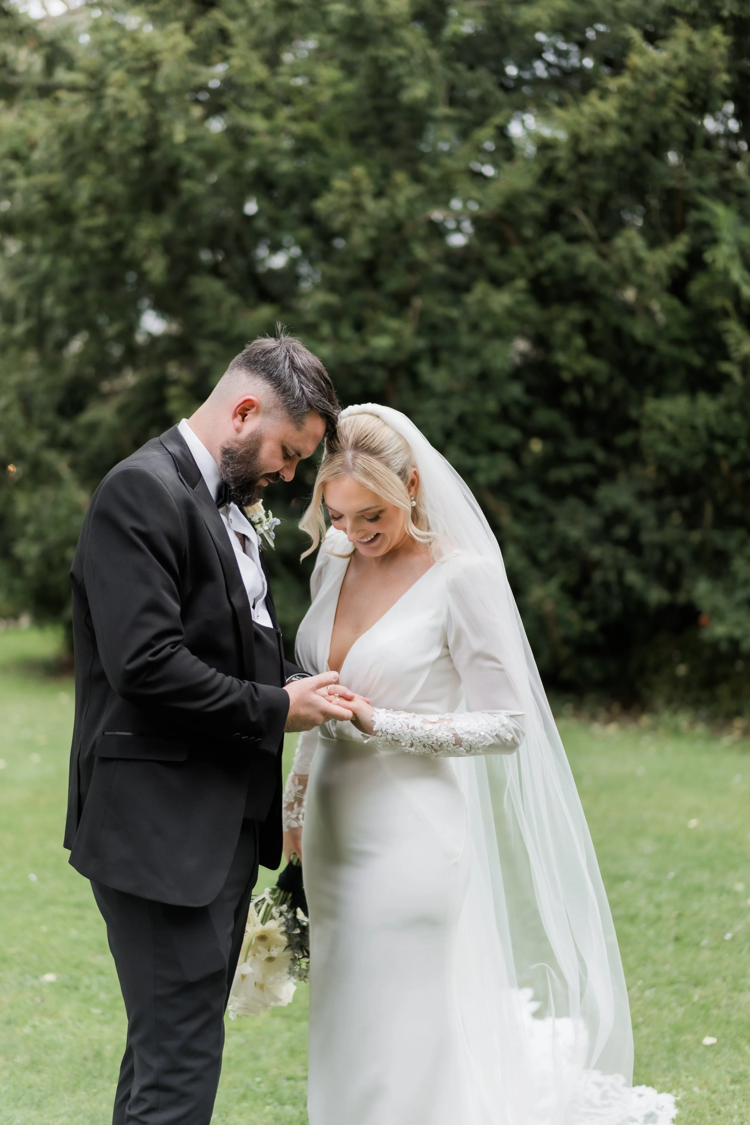 A bride and groom exchanging wedding rings outdoors, with a backdrop of green trees.