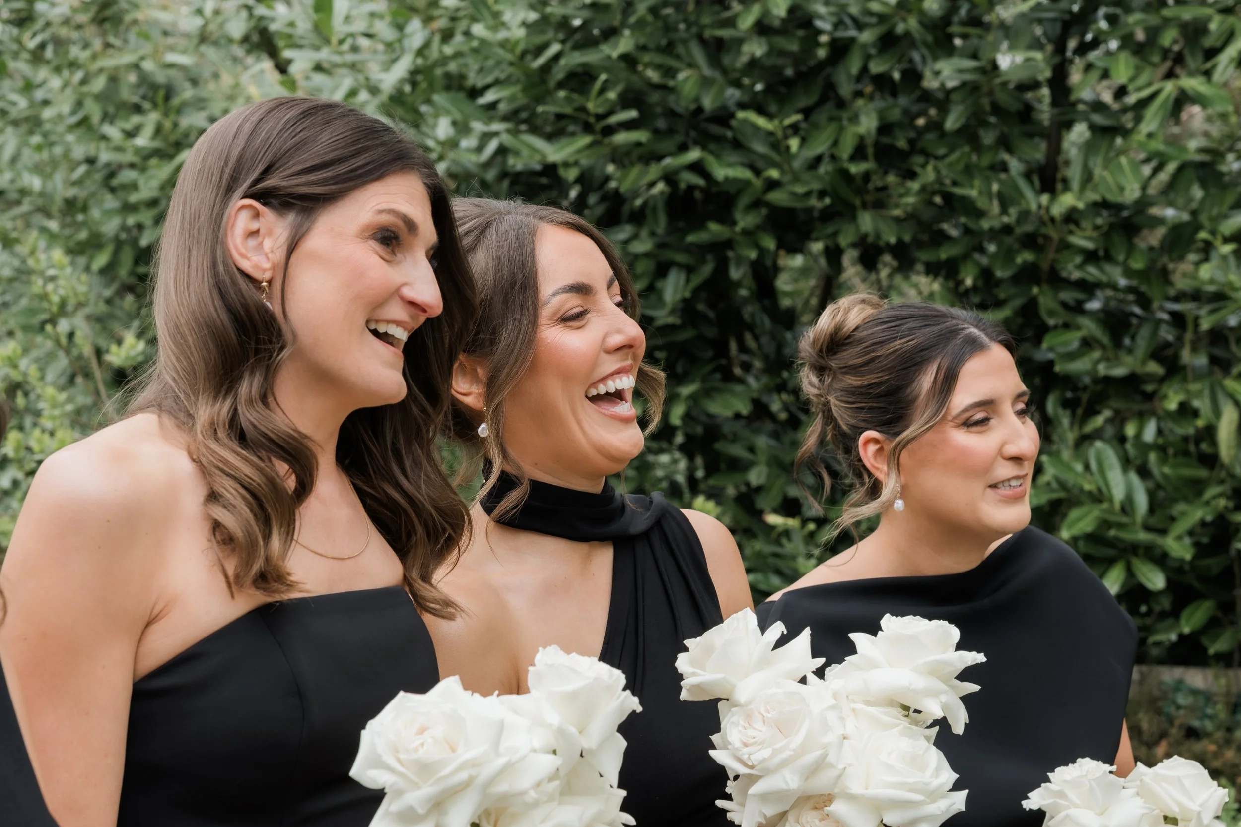 Three women dressed in black attire holding white floral bouquets, smiling and laughing outdoors with green foliage background.