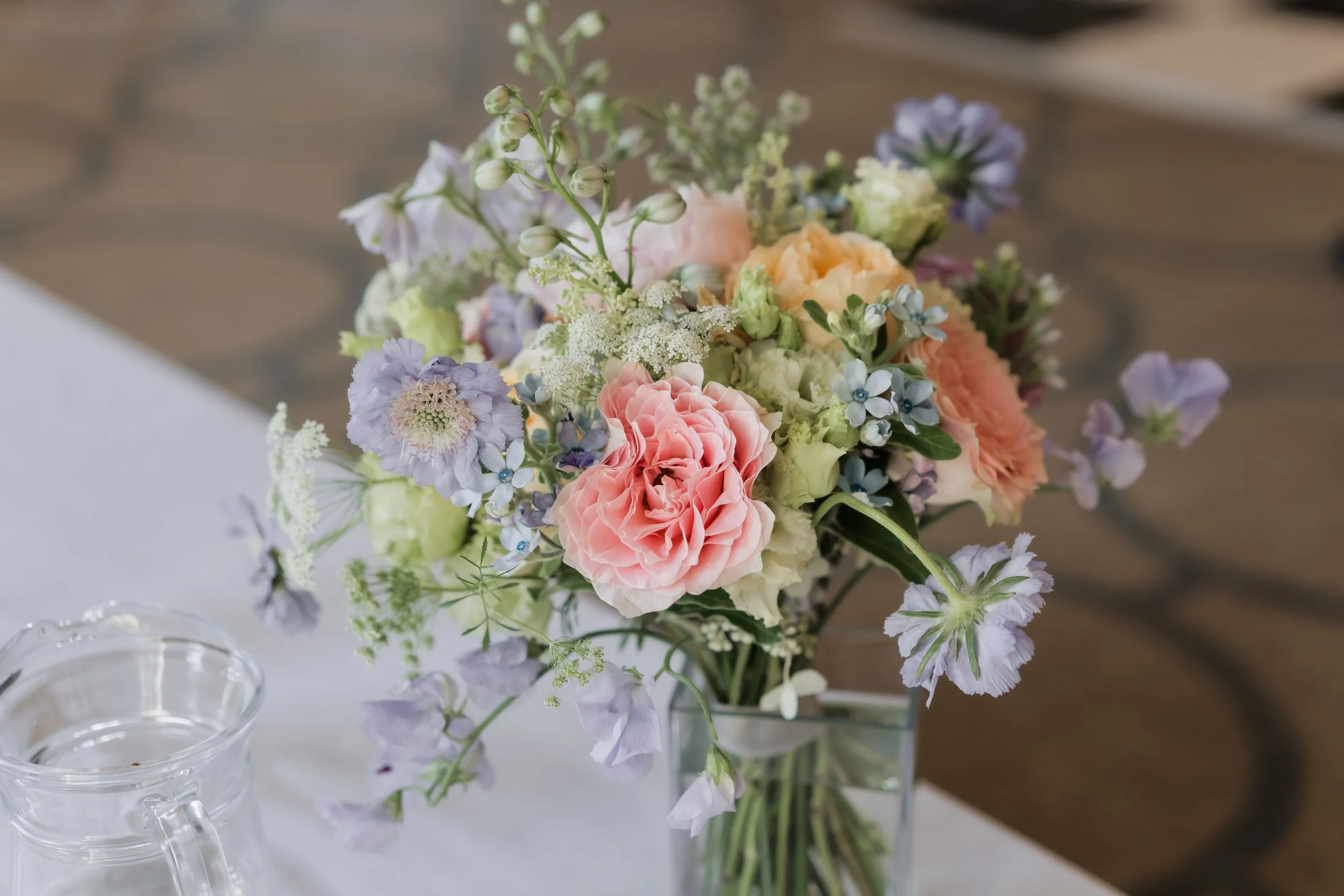 A bouquet of pastel-colored flowers including pink, purple, orange, and white, arranged in a clear glass vase on a table with a white tablecloth. There is a glass pitcher of water next to it.